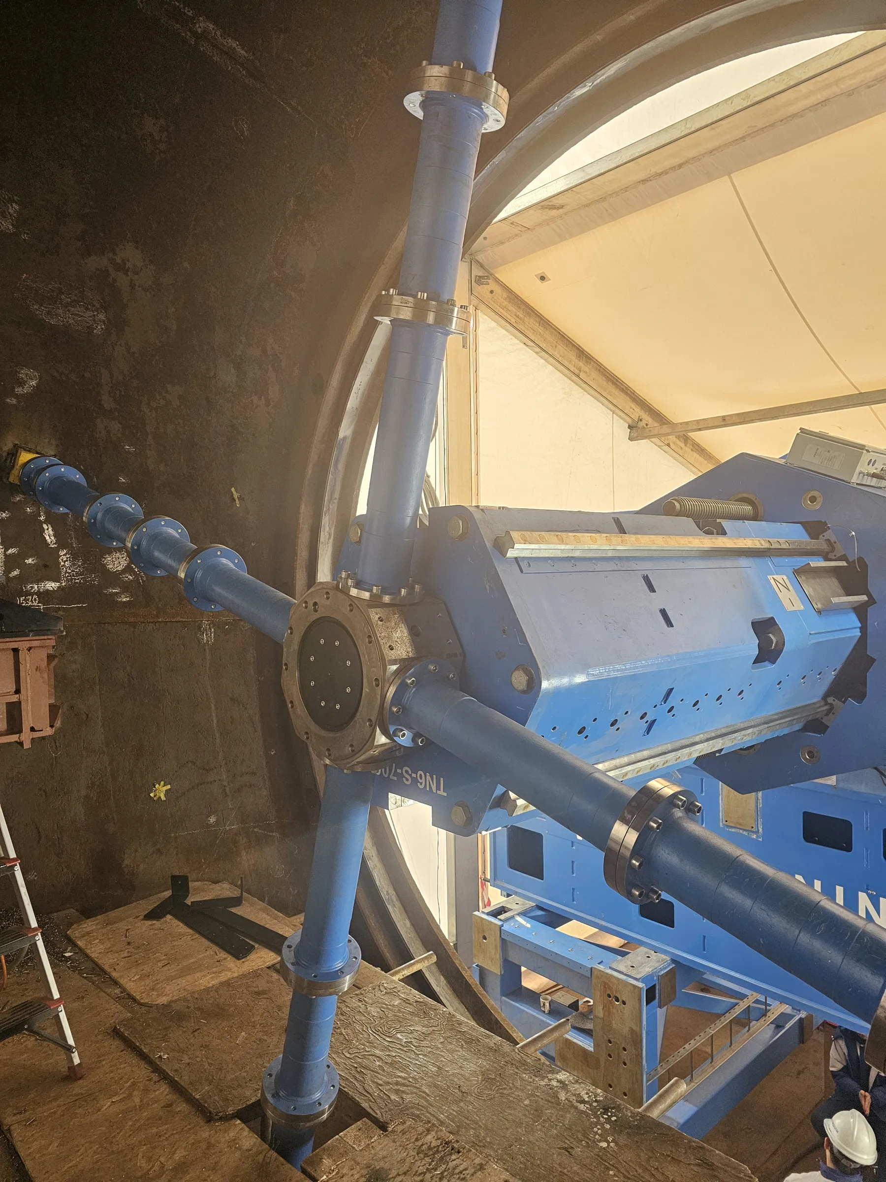 A large blue industrial machine is situated within a dark, cylindrical chamber, connected by blue pipes with metallic flanges. These pipes form a star-like configuration, with one extending along the chamber wall and others connecting to the machine. The floor consists of wooden panels, and a person in a white hard hat is partially visible. Bright, ambient light illuminates the scene from a light-colored enclosure on the right.