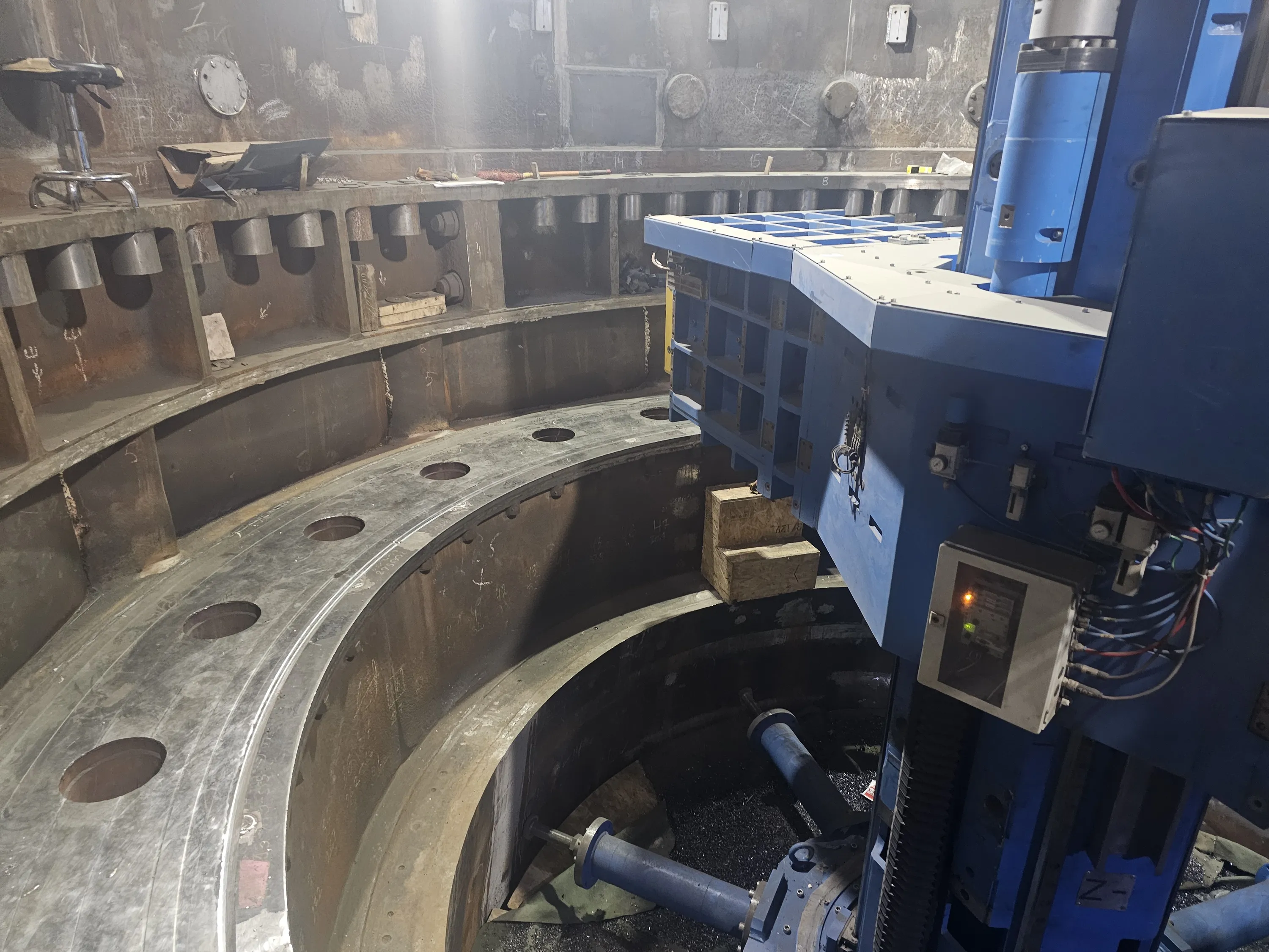 A large blue industrial boring machine is positioned over a massive, circular metal workpiece in a factory setting. The workpiece features multiple layers, numerous bolt holes, and a rough, unfinished surface. Below the machine, within the circular structure, are blue cylindrical components surrounded by metal shavings. In the background, a grey metal wall displays various markings, circular covers, and a ledge holding a stool and tools. Bright overhead lighting illuminates the scene, highlighting the metallic textures.
