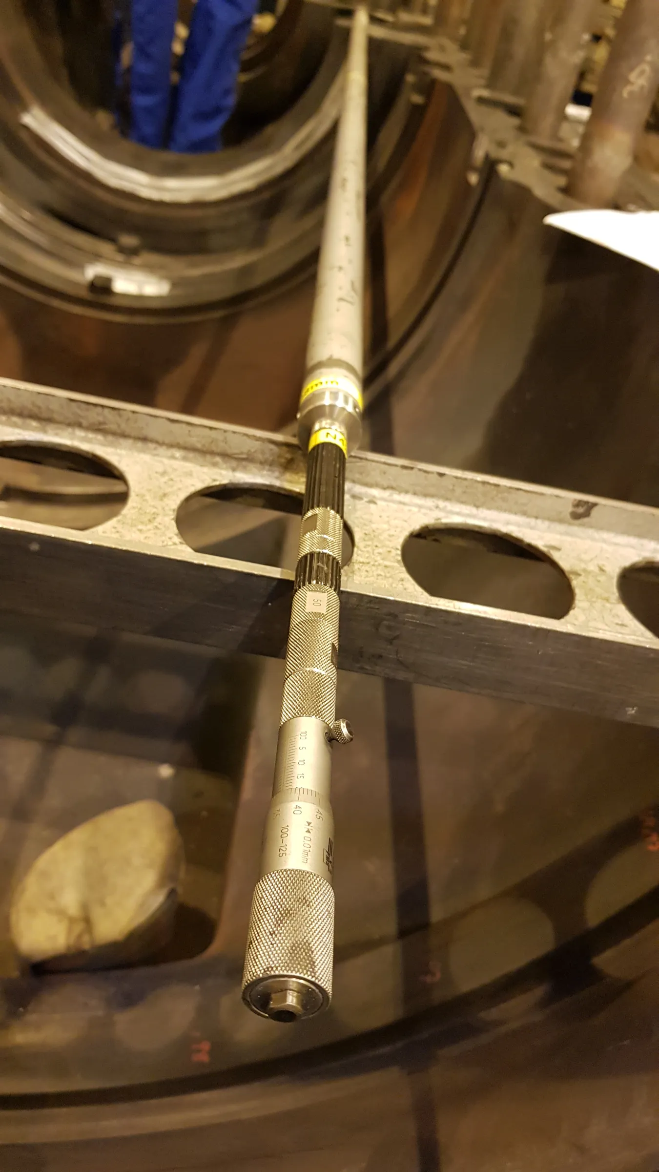 A long, silver and black internal micrometer tool is positioned vertically within a large, dark industrial assembly. The tool's knurled handle and scale, marked '100-125 0.01mm', are clearly visible. In the blurry background, a person in blue workwear is seen inside a circular opening, while a grey metal frame with oval cutouts spans the mid-ground. Dim lighting creates reflections on the metallic surfaces.
