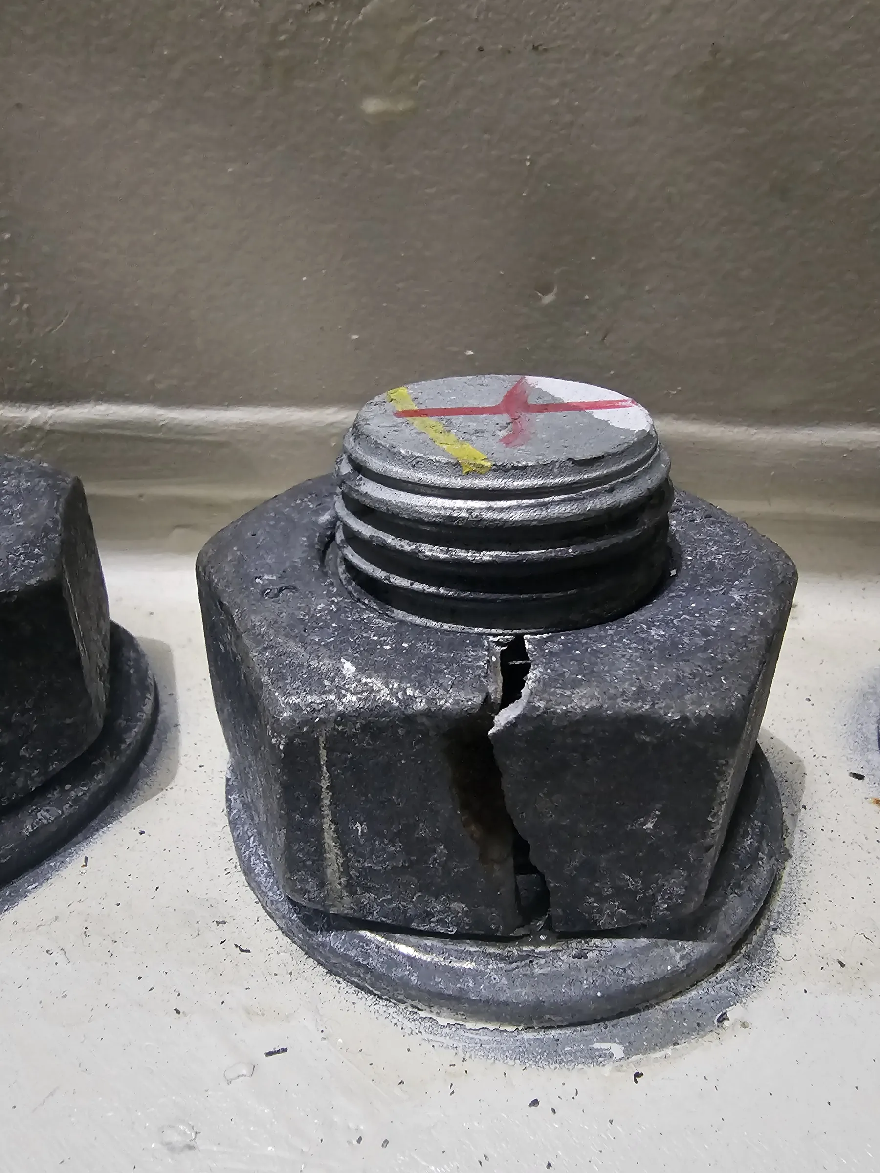 A close-up view features a large, dark grey hexagonal nut with a significant vertical fracture on one side. A threaded bolt head, marked with red and yellow paint forming a cross on its flat top, extends from the nut. A second, similar nut is partially visible to the left. These industrial fasteners are set on a light-colored base, against a light grey background. Bright, even lighting illuminates the textured surfaces.