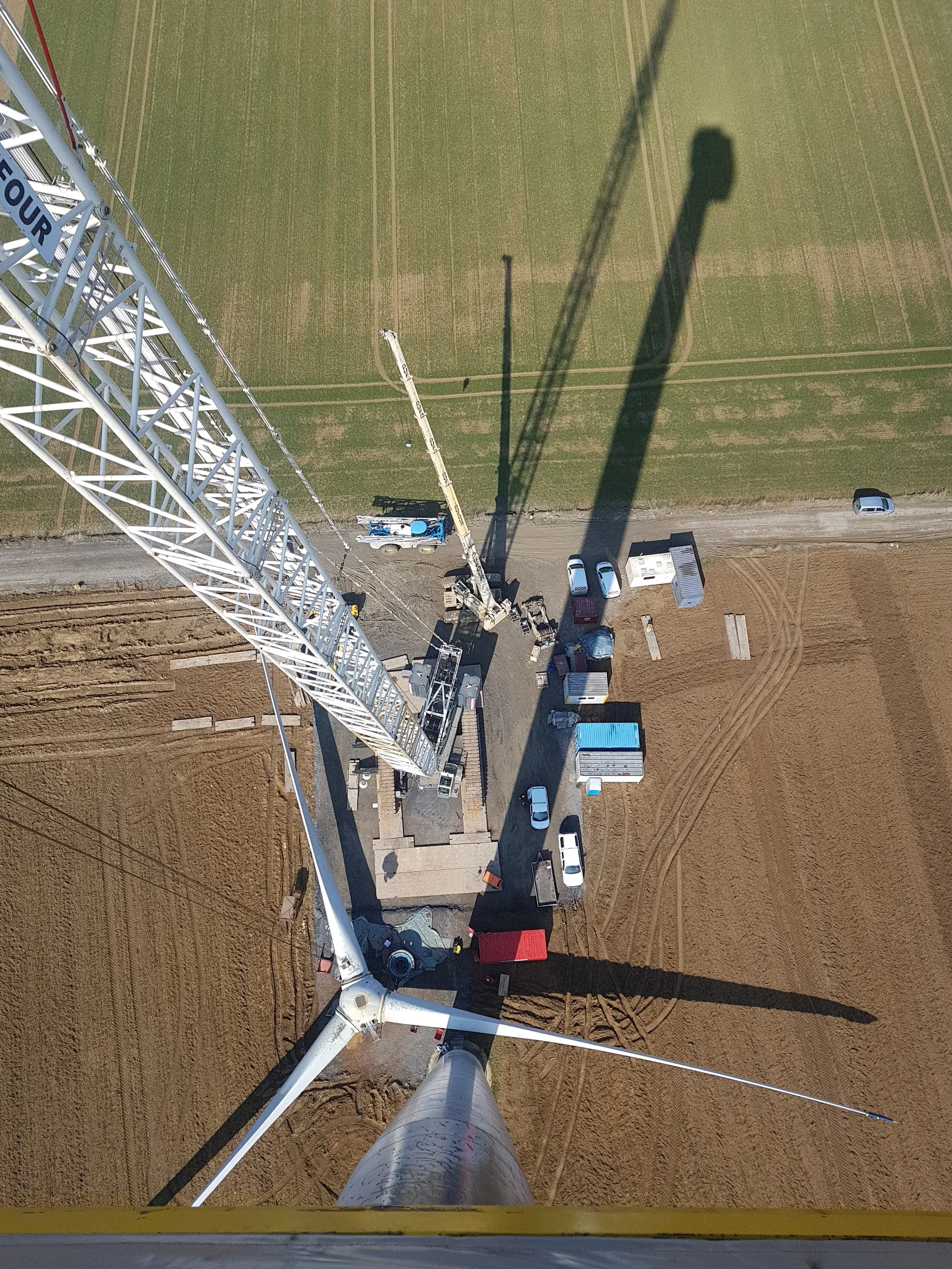 A high-angle aerial view shows a wind turbine construction site under bright sunlight. A large white lattice crane boom extends from the upper left, while a silver turbine tower with three white blades rises from the bottom center. The ground features brown, tilled earth with tire tracks, a dirt road, and a green field in the background. Various construction vehicles, equipment, and containers are visible, casting long, distinct shadows.