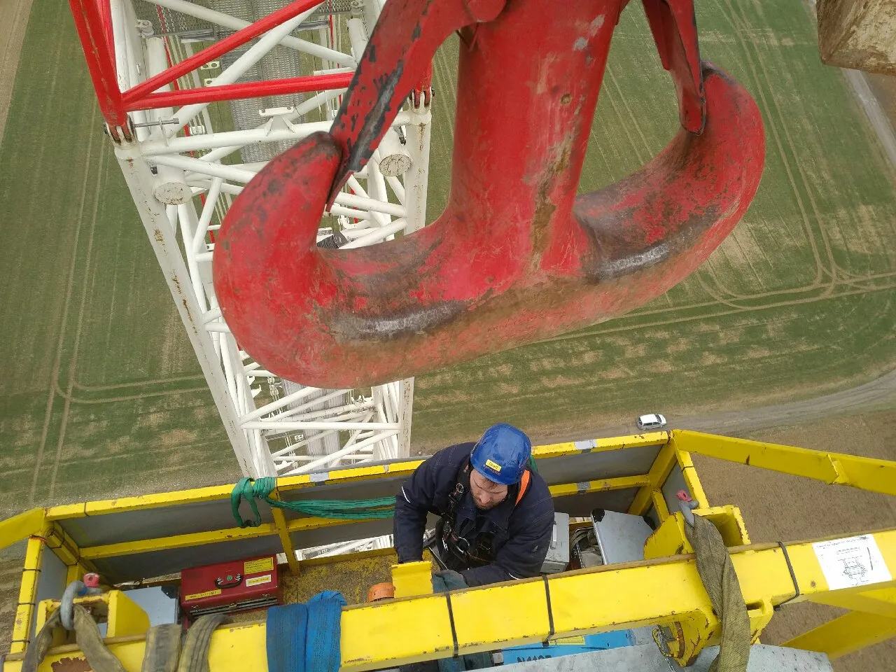A high-angle view captures a large, worn red crane hook in the upper frame, with a white lattice crane structure extending below it. In the foreground, a worker in a blue hard hat and dark attire stands within a yellow elevated work platform, which contains various straps and a red control box. The background features a wide green field marked with faint tracks, and a small white vehicle is visible on a distant path. The scene is brightly lit by natural daylight.