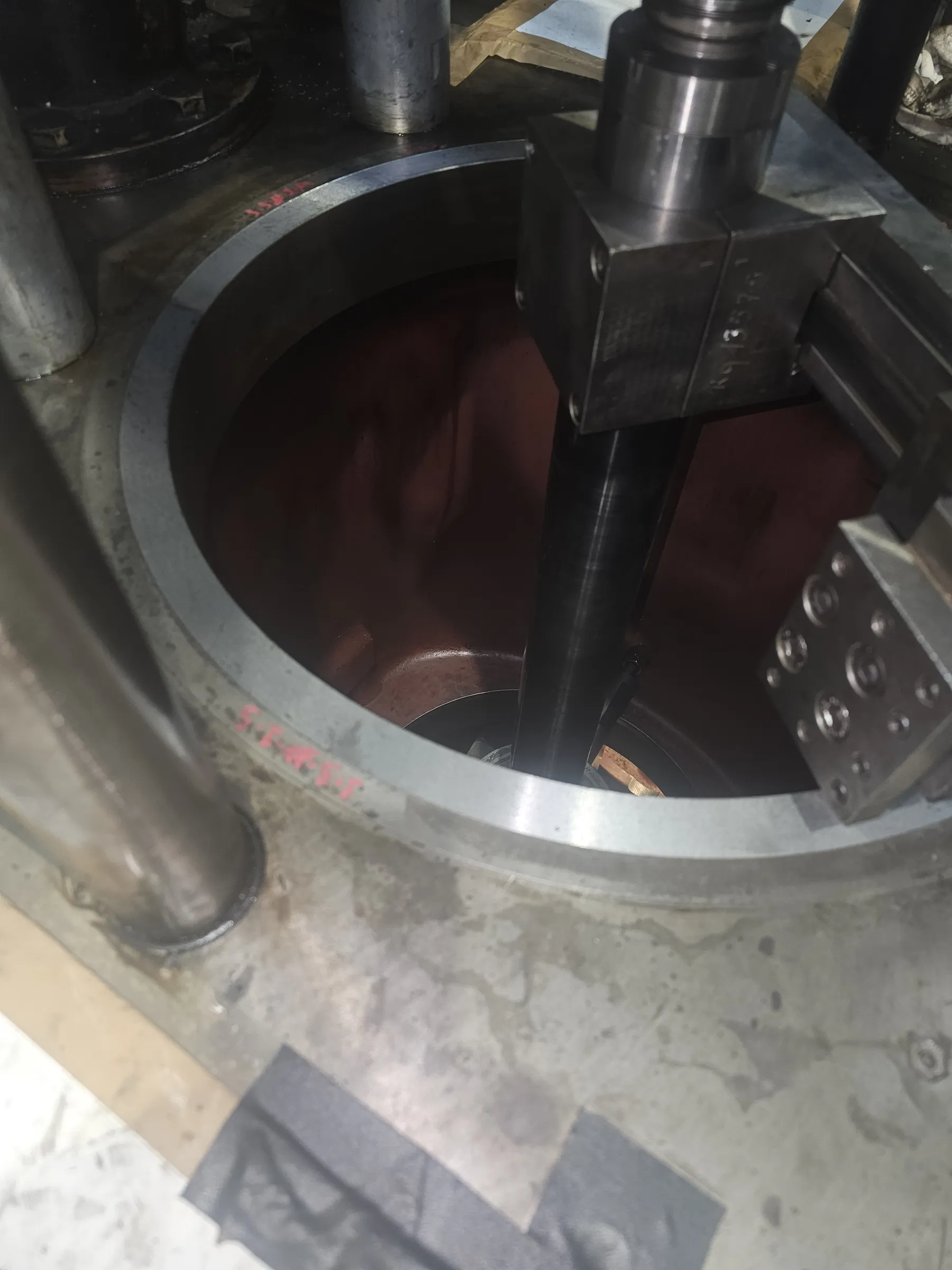 A top-down view shows a large, dull grey metal cylinder opening with red painted markings on its rim. The cylinder's interior is a dark reddish-brown. A dark metal shaft descends into the cylinder from a bolted metal assembly on the upper right, which has etched numbers. The surrounding surface is a lighter grey with some liquid marks.