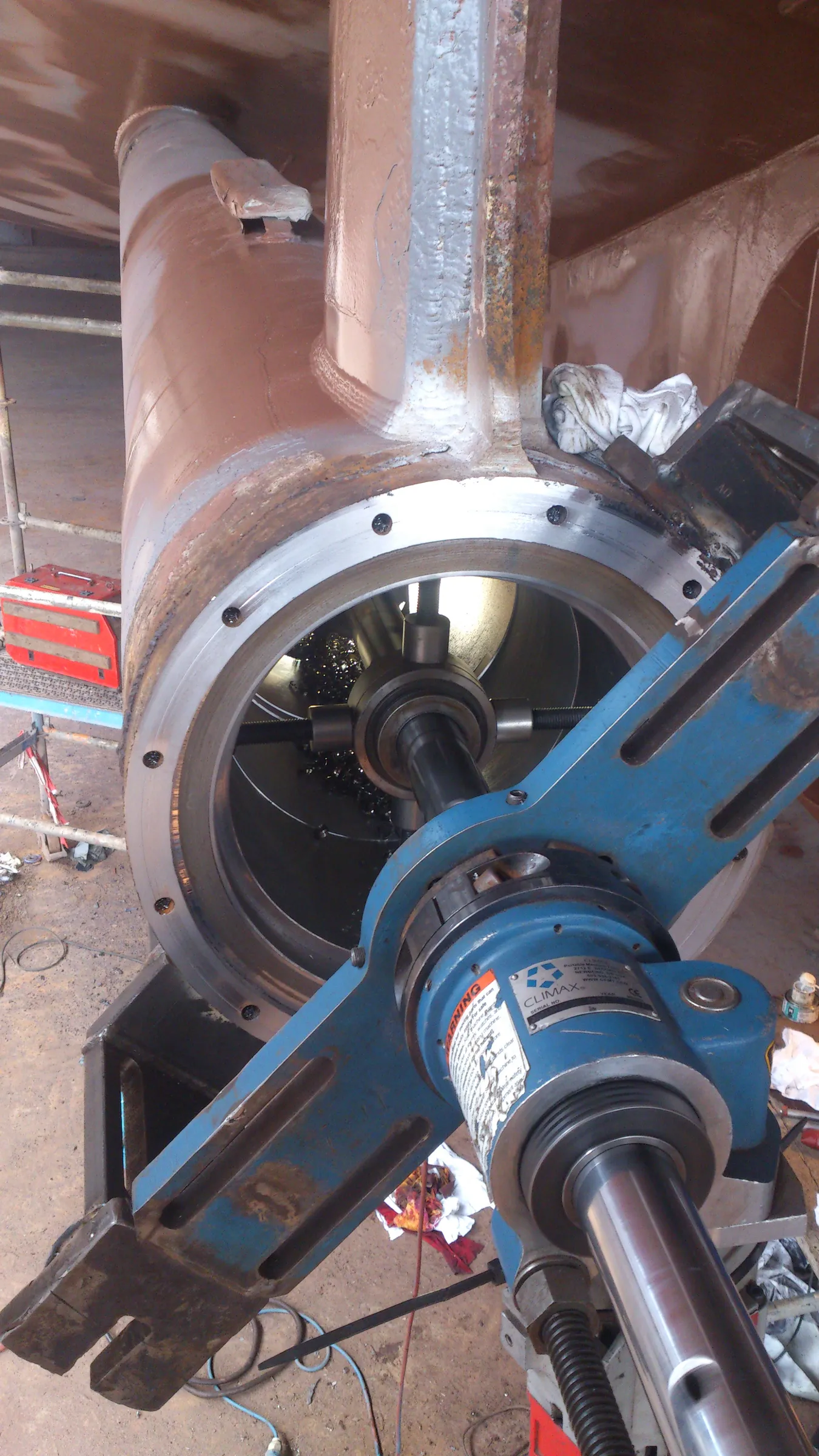 A large, rusty brown cylindrical metal structure is being machined by a blue portable line boring tool. The tool's shaft extends into the cylinder, where dark metal shavings are visible from the ongoing work. Scaffolding and a red toolbox are on the left, while a white rag rests on the structure to the right. The scene depicts an industrial repair or fabrication process under bright lighting.