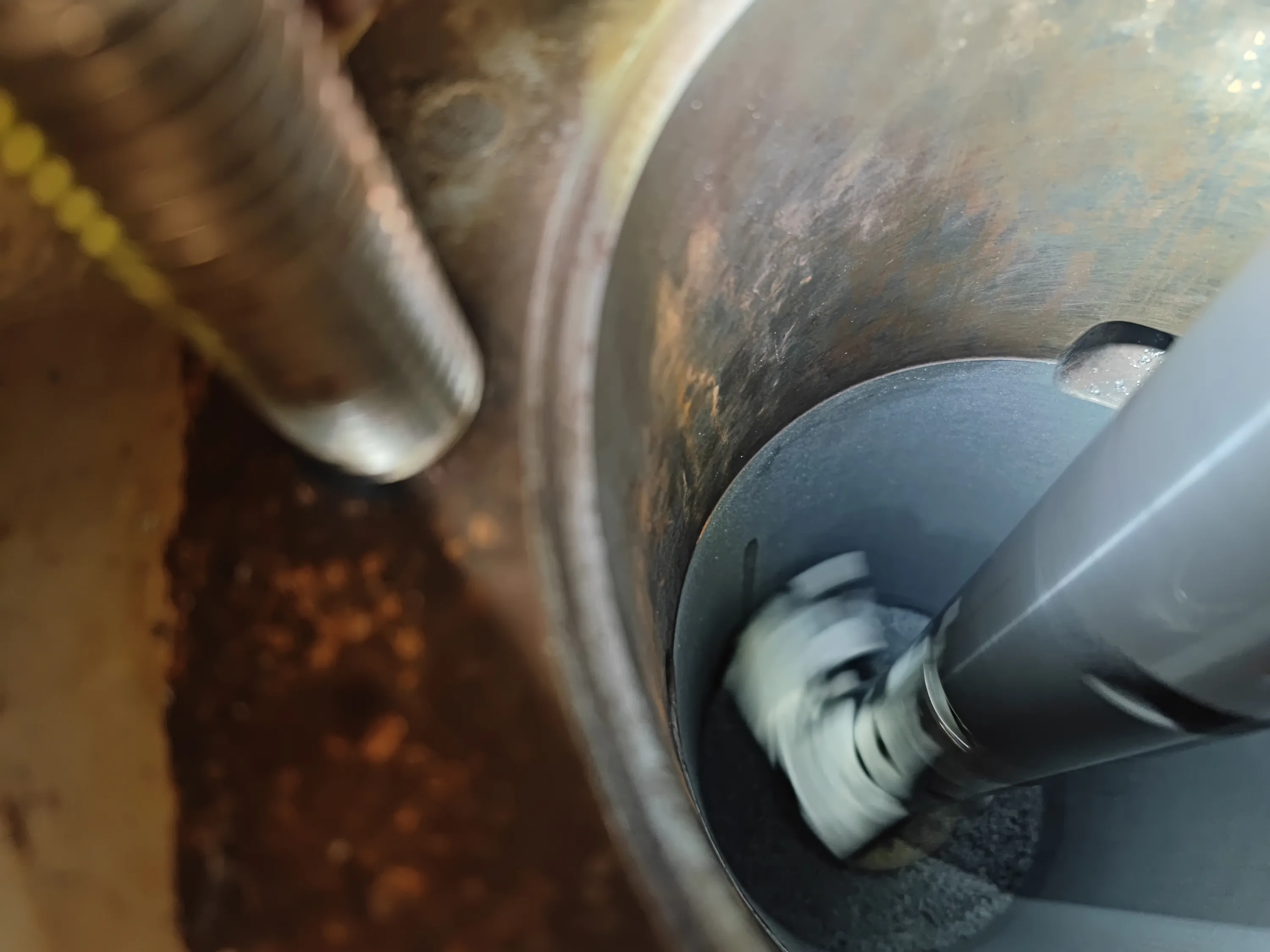 A rotating brush tool cleans the dark, carbonized interior of a metal cylinder, with a threaded rod and yellow cable visible nearby. The cylinder's upper walls show rusty brown textures.