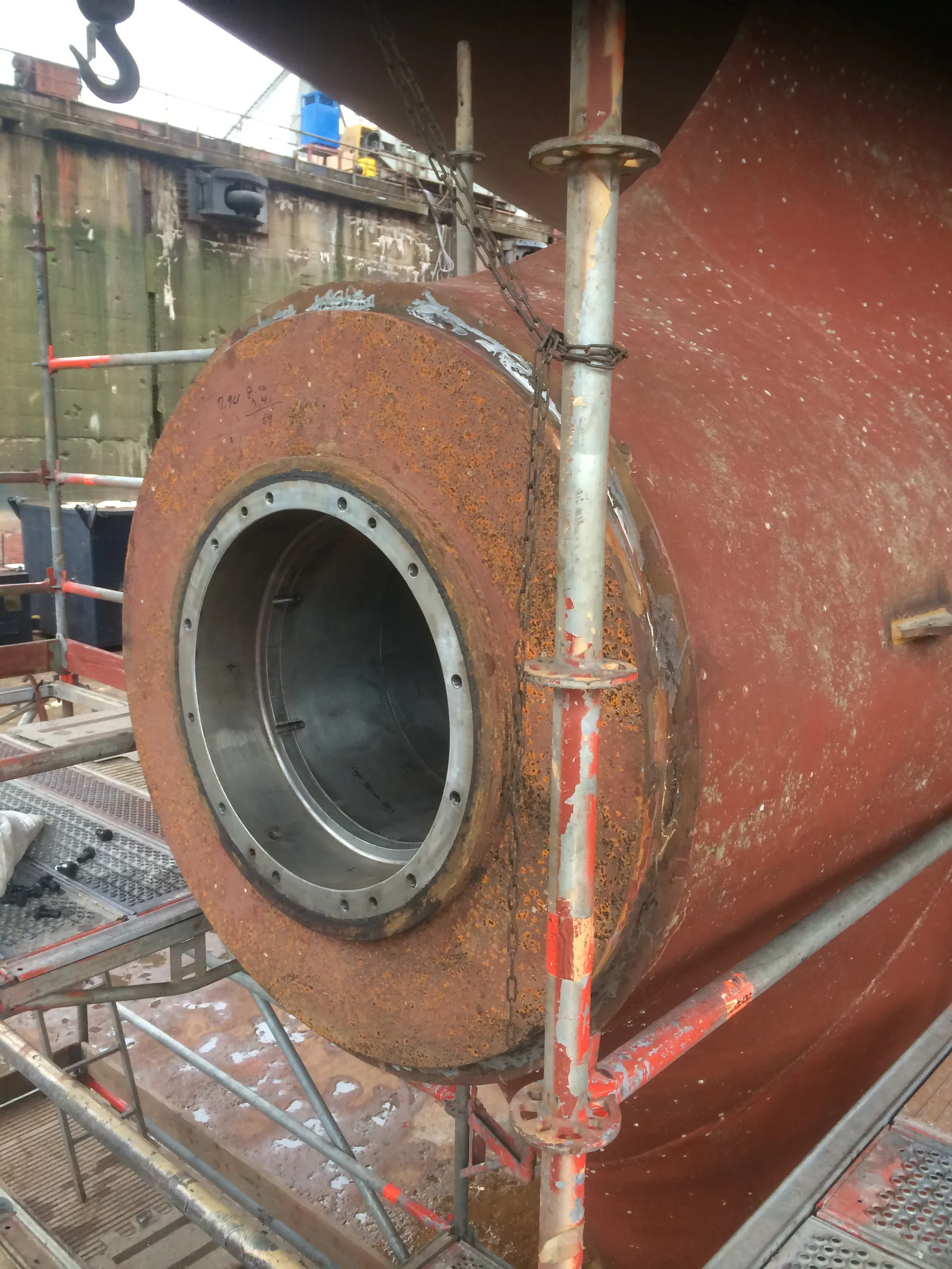 A large, rusty cylindrical metal structure, possibly a stern tube, is seen in a dry dock, supported by silver and red-painted scaffolding. Its clean inner ring has multiple bolt holes. A red ship hull is visible to the right, and a concrete dock wall with a black bollard and industrial equipment is in the background under diffused light.