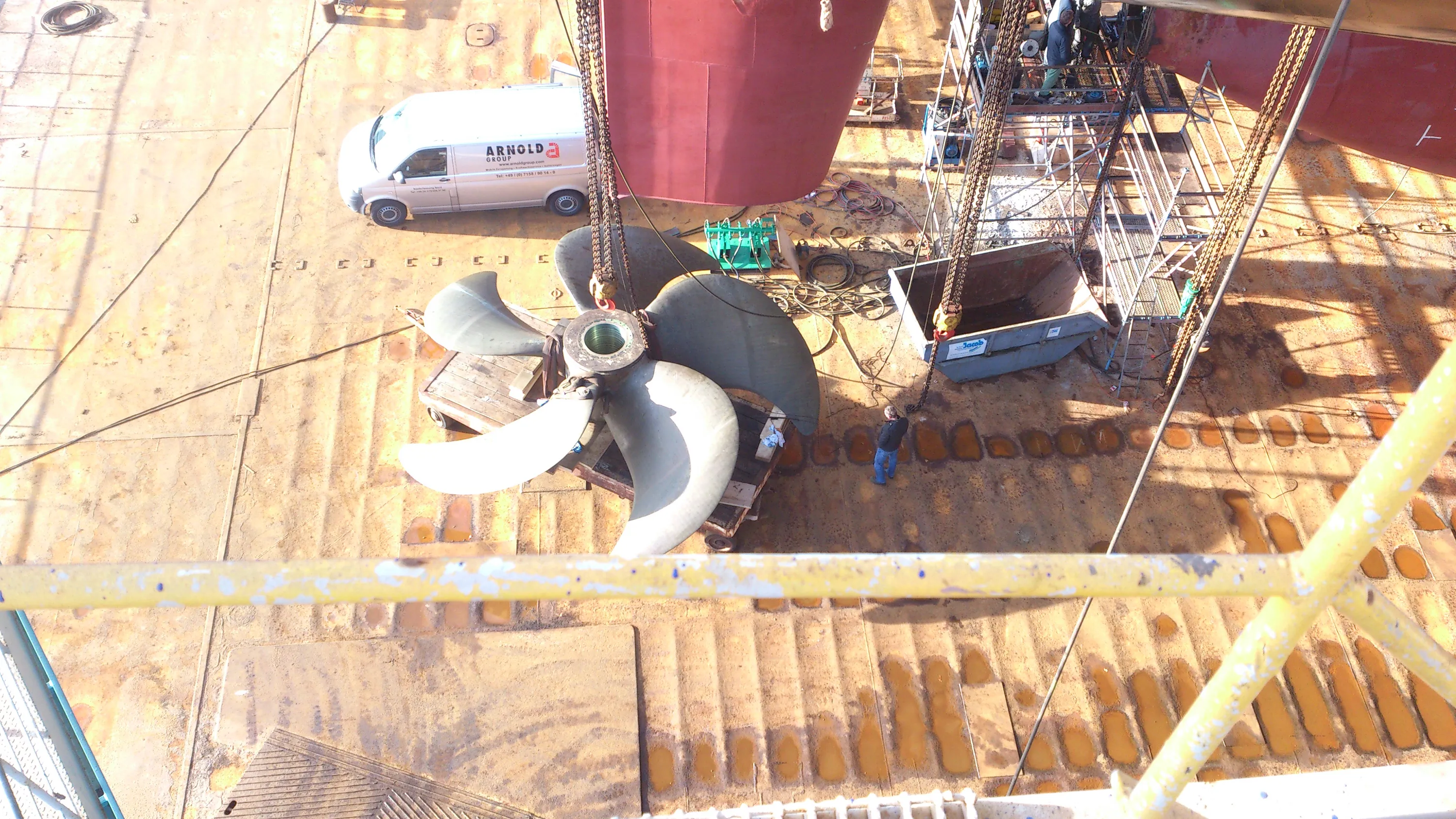 An overhead view shows a large ship propeller resting on a wooden base on a rusty dry dock floor. A white service van is parked near the red hull of a ship, with scaffolding and a metal skip being lowered by chains. A worker stands on the ground, and another is visible on the scaffolding. The metal ground has darker, wet-looking patches, all illuminated by bright sunlight casting strong shadows. A yellow railing is in the foreground.