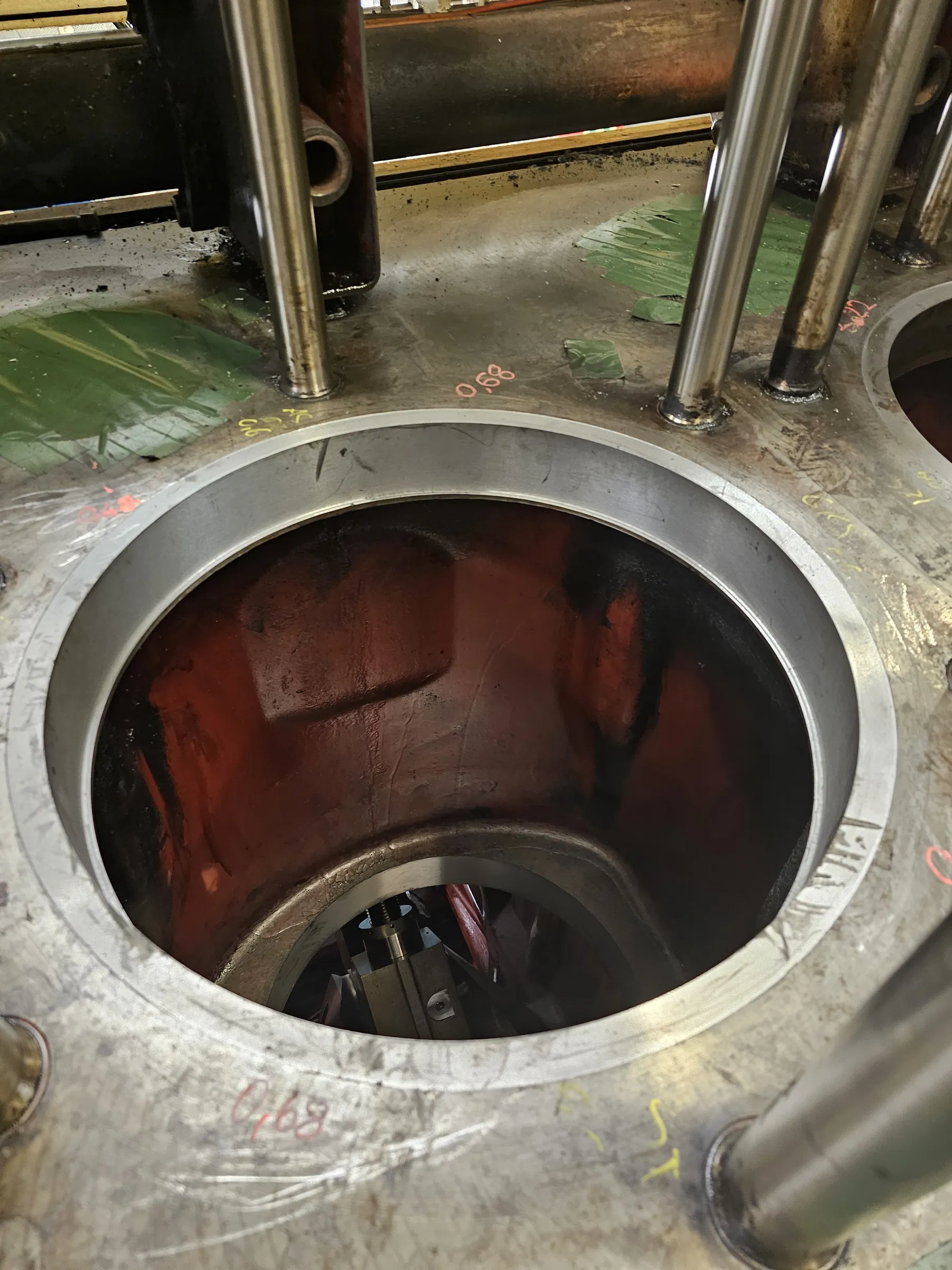 A high-angle shot captures the open cylinder bore of a large engine or industrial machine. The metallic grey surface of the engine block surrounds the dark, reddish-brown interior of the cylinder, where mechanical components are faintly visible at the bottom. Several shiny metal rods extend vertically from the block. Pink and yellow numerical and alphabetical markings are visible on the block's surface, along with scattered green material and dark stains on the surrounding floor.