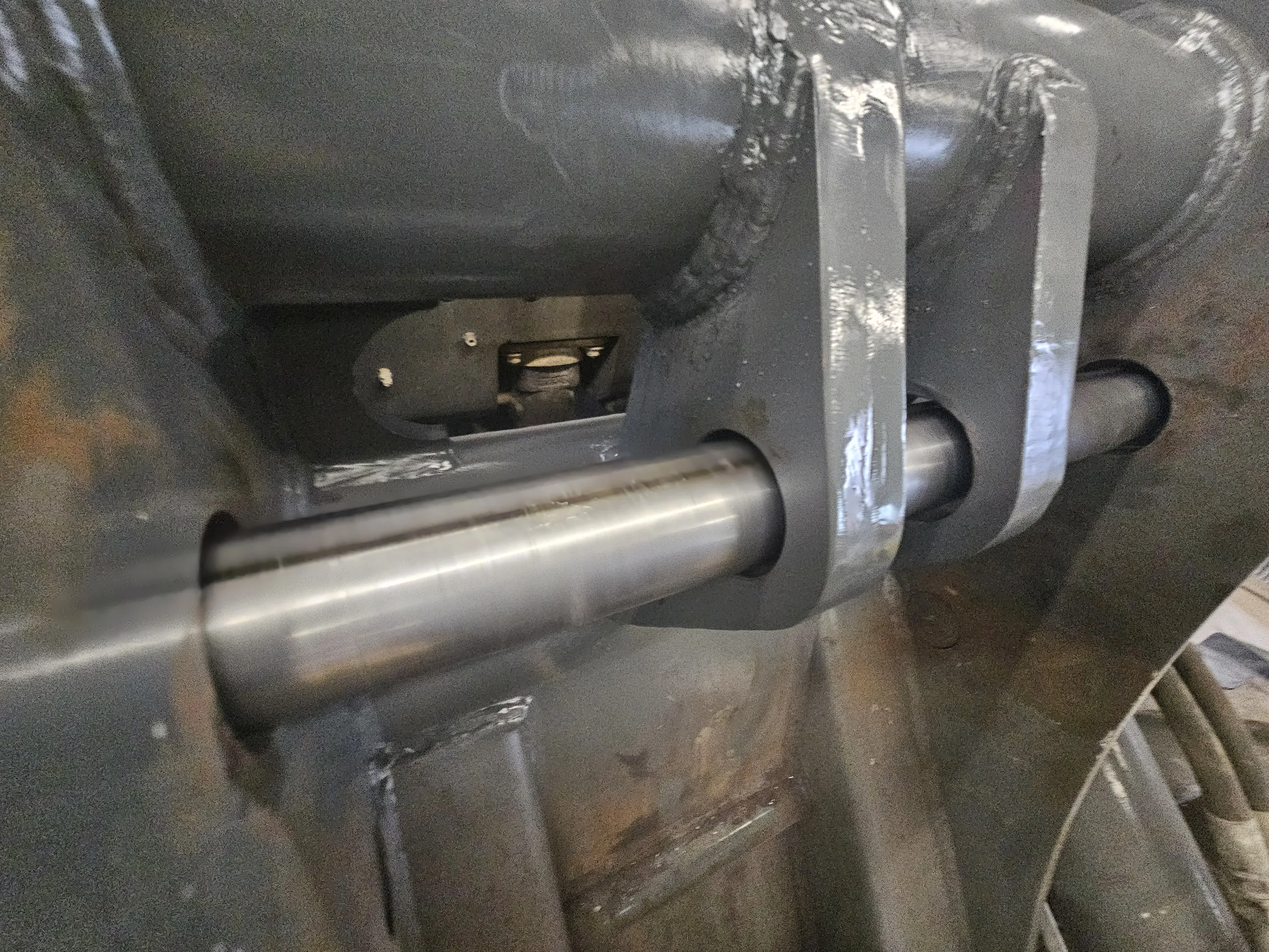 A close-up shot of heavy machinery components, featuring a polished silver metal shaft passing through a dark grey, robust metal clevis bracket. The bracket is welded to a larger, textured grey frame. Light reflects off the shaft and welded seams, illuminating internal mechanical elements visible in the background.