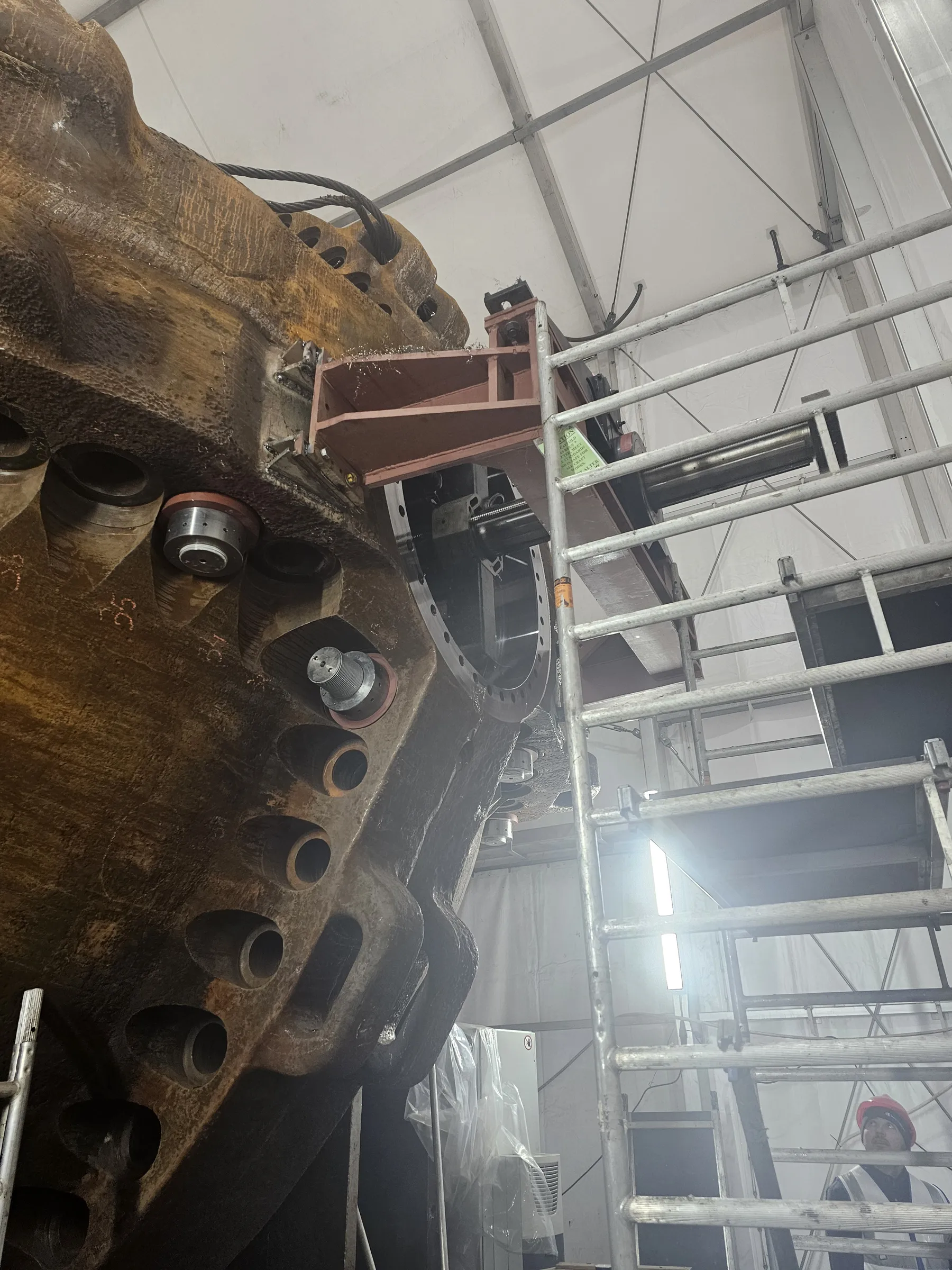 A massive, textured, rust-colored industrial component with various openings and fittings occupies the left side of the image. A fabricated metal bracket, covered in metal shavings, is attached to the component. On the right, silver scaffolding rises towards a white tented ceiling supported by metal trusses. Bright artificial lighting from the right creates significant glare. A worker wearing a red hard hat and a high-visibility vest is visible behind the scaffolding at the bottom right.