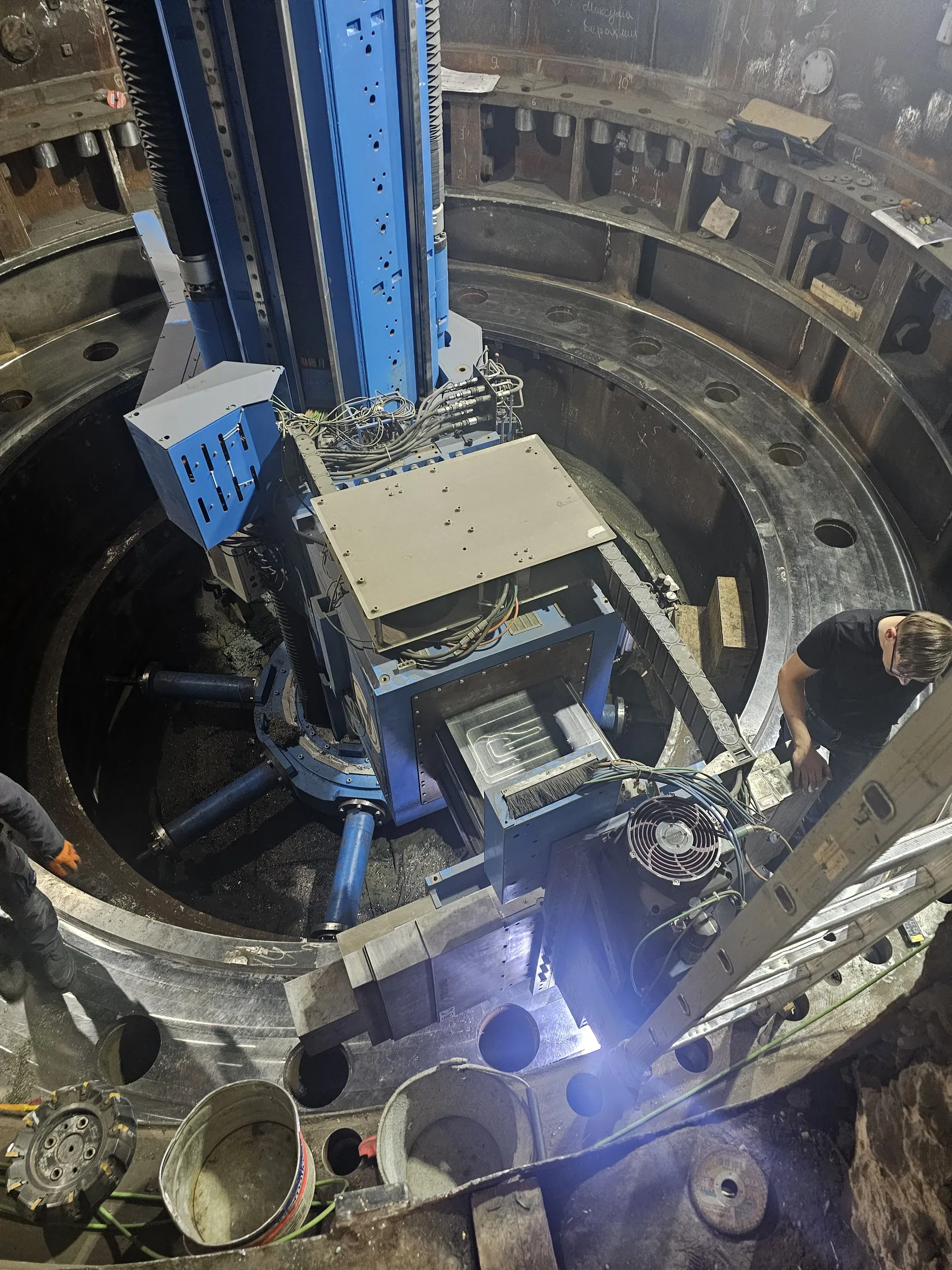 A large blue and grey industrial machining tool is positioned inside a massive circular metal structure, actively working on its inner surface. A worker in a black shirt and glasses observes the operation from the right, near a ladder, while another worker's legs are visible on the bottom left. The machine features a central base with three support arms, a vertical column, and a horizontal arm with a cutting head. Various cables, a fan, and metal buckets are also present. Bright spotlights illuminate the metallic surfaces, creating strong reflections and highlighting the work area.