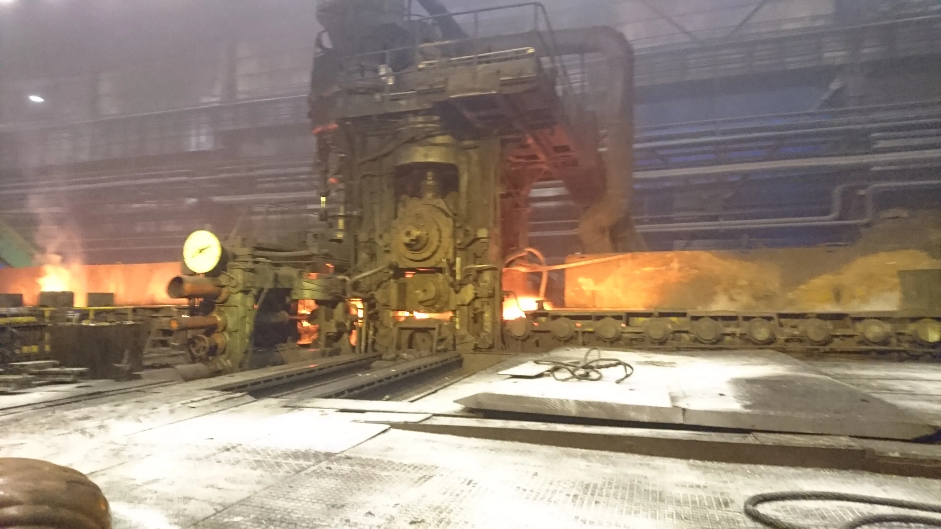 An industrial facility features a large, complex metal processing machine with gears and pipes, illuminated by the intense orange glow of hot metal. To the right, a long conveyor system carries a massive, incandescent orange slab. The foreground shows a light gray, dusty floor with metal plates and coiled dark cables, while the background is filled with pipes and structures under a hazy ceiling.
