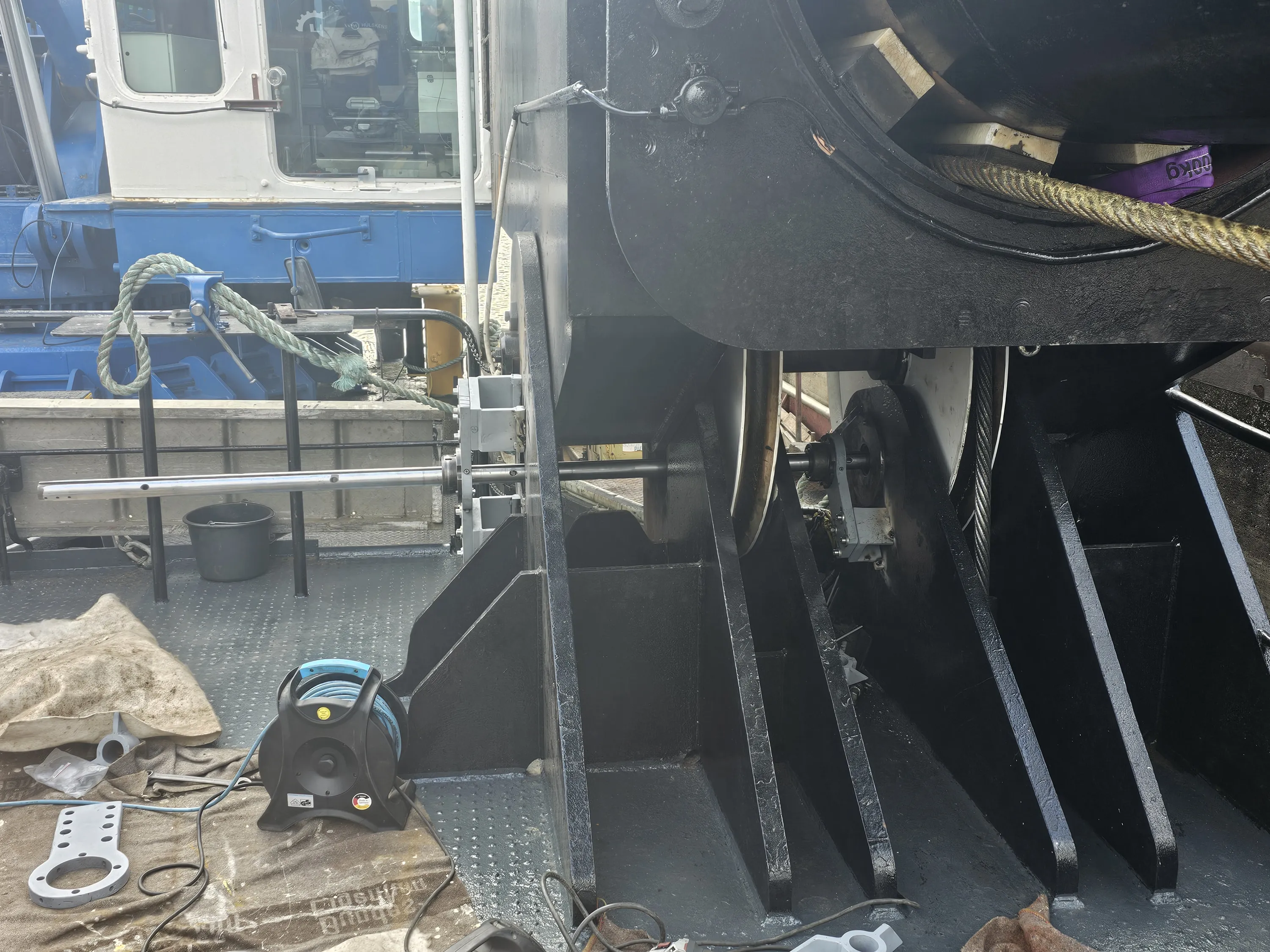 A bright outdoor scene captures industrial equipment on a vessel deck. A white and blue cabin structure is visible on the left, with a thick green rope secured to a blue cleat. The dark grey textured deck holds a black and blue hose reel, grey metal parts, and a dark bucket. The right side features large black machinery, likely a winch, with light-colored pulleys, a thick metal wire rope, and exposed electrical wiring. A purple lifting strap marked "600kg" is visible within the mechanism.