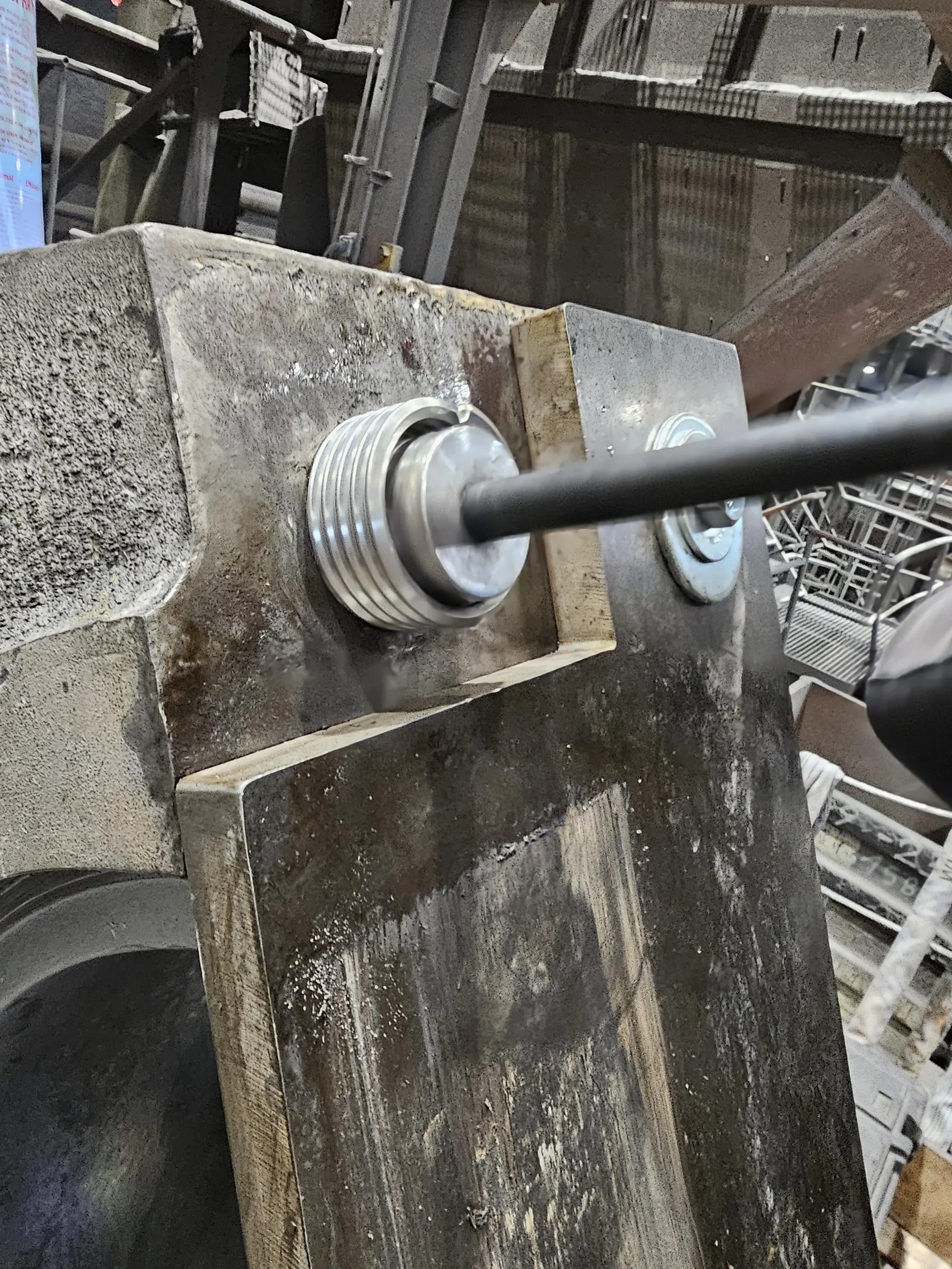 A close-up view captures heavy industrial metal components, with a textured grey block on the left and a smoother dark plate on the right. A thick, dark rod extends through both, secured by a series of shiny metallic rings and a flat washer. The surfaces appear grimy and worn, with some reflective wet areas. The background shows blurred metal structures and scaffolding under dim lighting.