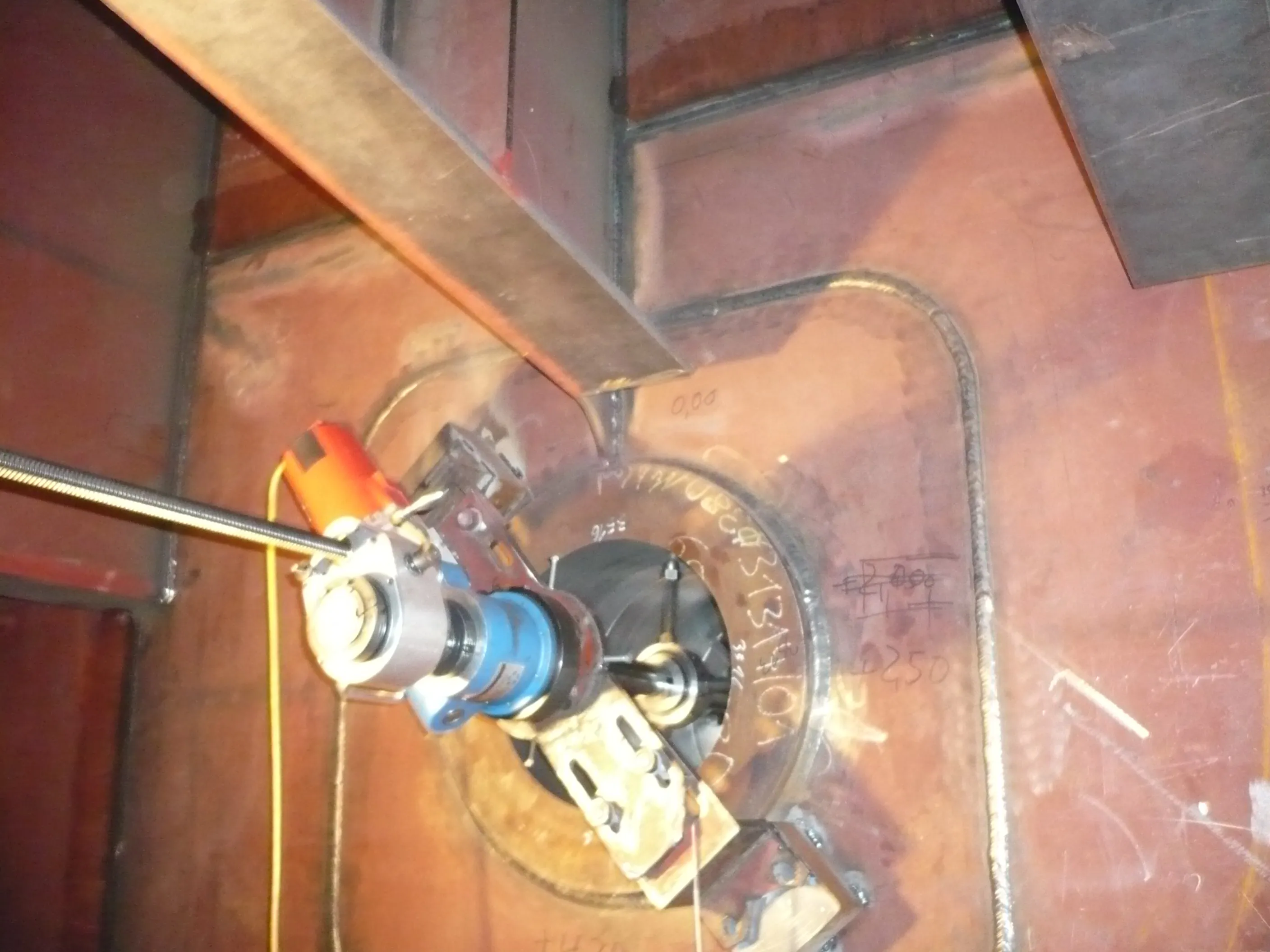 An industrial line boring machine is positioned within a reddish-brown metal structure, likely a vessel or tank interior. The machine features a central shaft, blue and orange components, and is secured by metal brackets. Visible welds and handwritten numbers mark the steel plates, illuminated by artificial light.