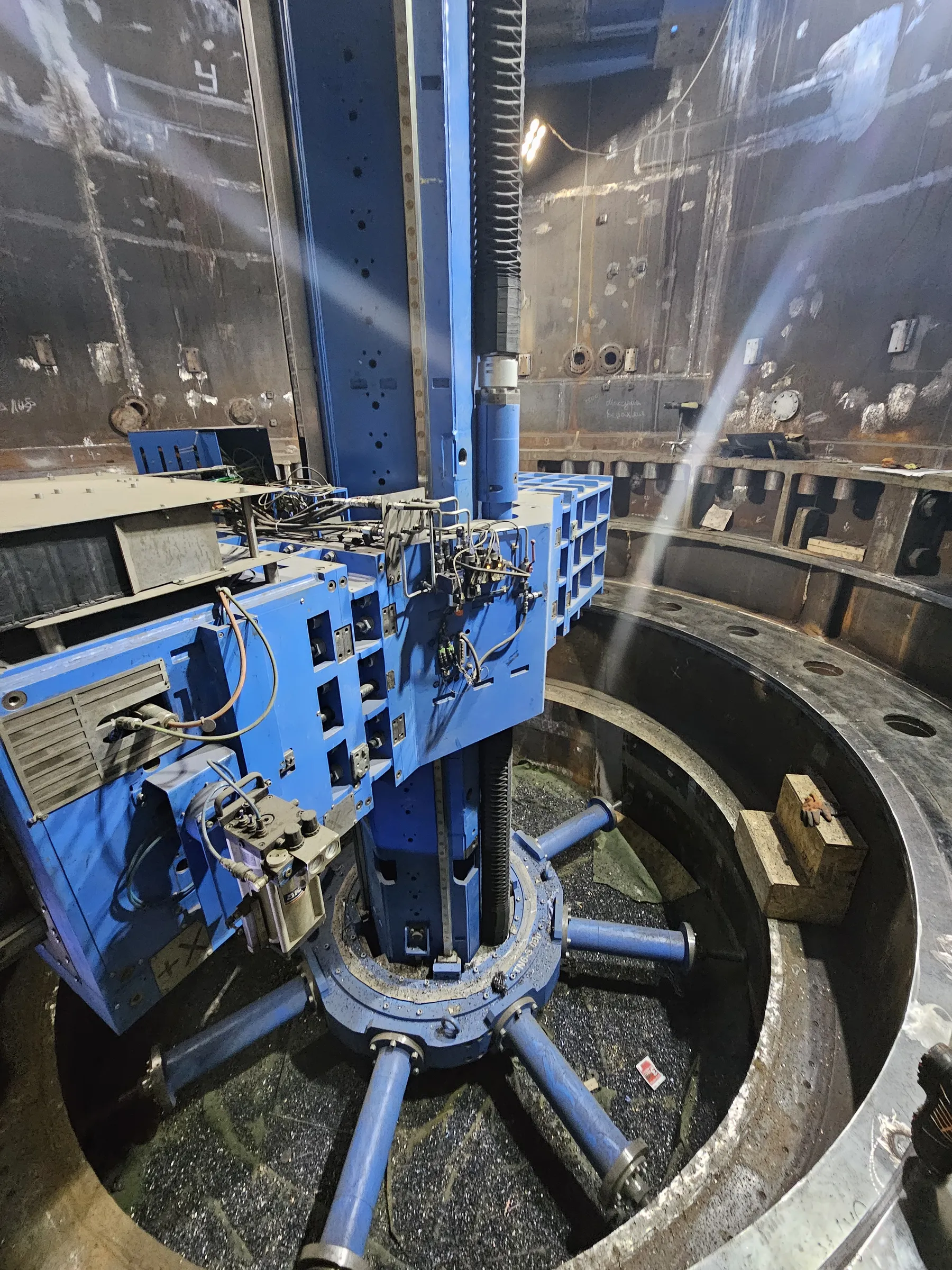 A large, blue industrial boring machine is positioned centrally within a massive, circular metal structure. The machine has a prominent vertical column with a bellows-like cover and a horizontal arm equipped with various controls, hoses, and wiring. Its base features several blue cylindrical supports extending into a floor covered with dark metal shavings. The surrounding raw metal walls of the structure are dark, showing visible welds, markings, and some surface grime, illuminated by bright industrial lights casting rays.