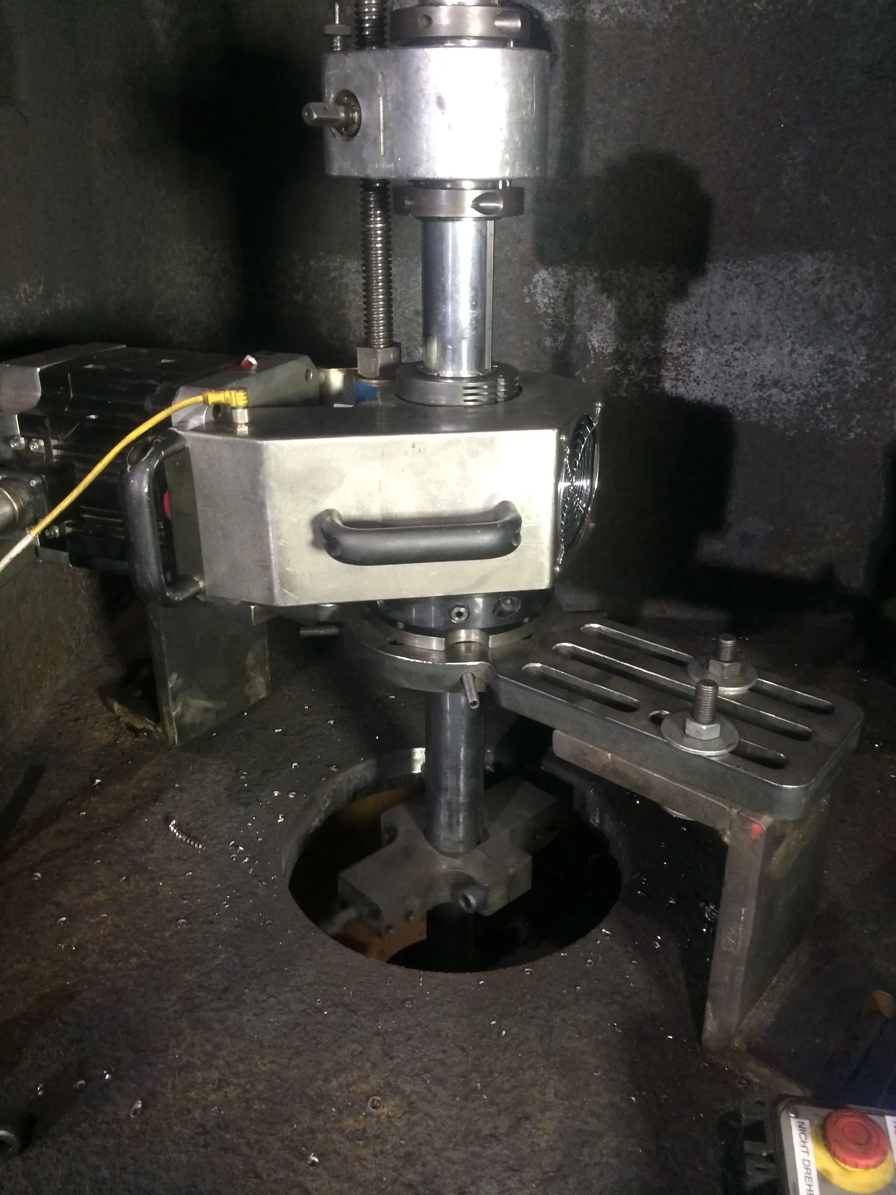A dimly lit view of industrial machinery, centered on a vertical assembly of silver metal components. This includes a cylindrical part at the top and a larger rectangular housing below, which features a black handle, a yellow cable, and a fan grill. A metal shaft extends from the housing into a circular opening in a dark, textured machine bed, connecting to a cross-shaped attachment. A bolted metal bracket is visible to the right, and a control panel with red and yellow buttons is partially seen at the bottom right. The dark machine surface is speckled with small, bright particles.