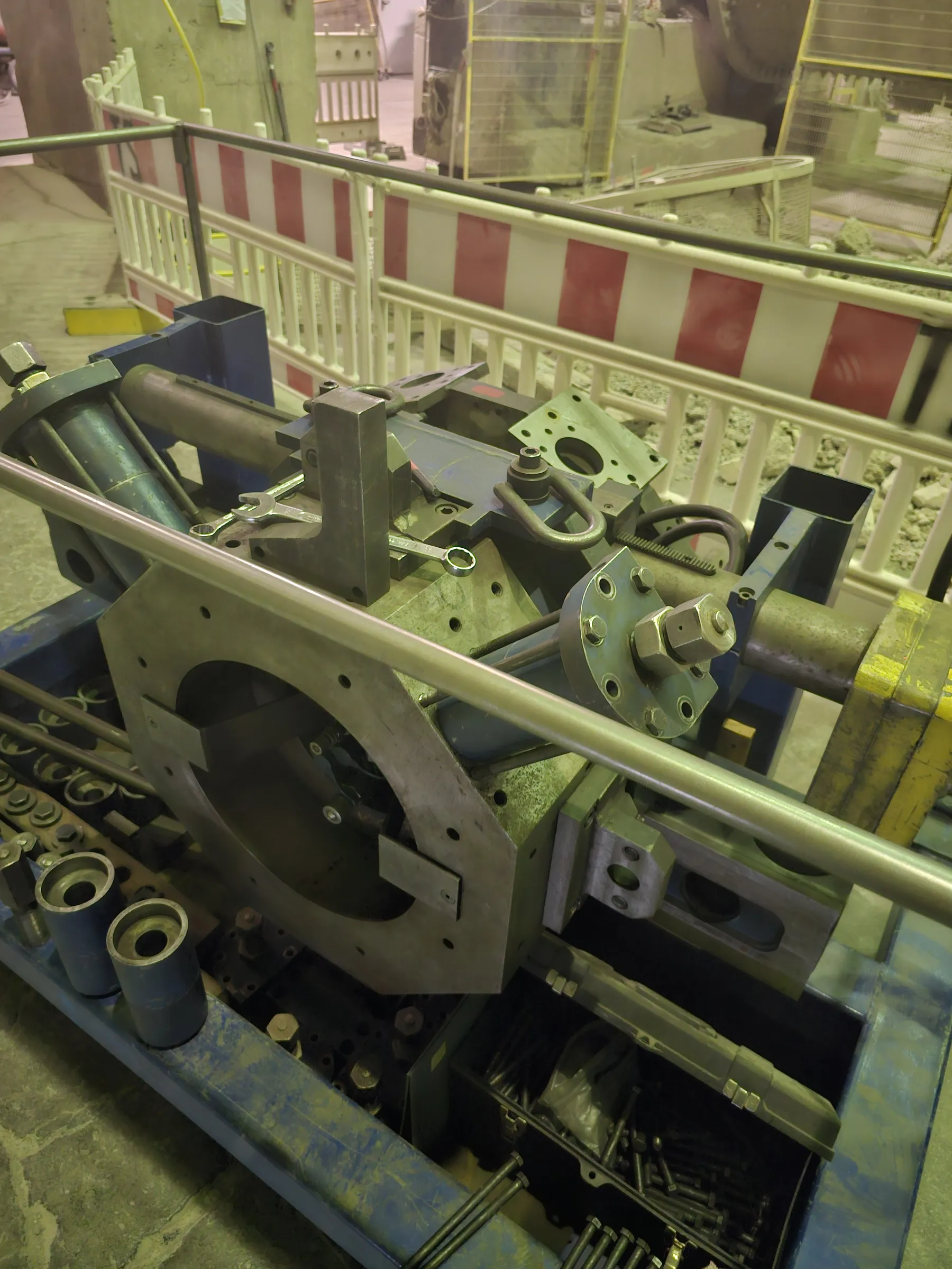 A robust industrial machine, featuring a blue frame and numerous grey metal components, is captured from an overhead angle. Prominent elements include large circular plates, cylindrical parts, and a long horizontal metal bar. Wrenches are visible on a central section, alongside a large yellow block. In the lower left, two blue cylindrical pieces rest on the frame, while a compartment in the lower right is filled with bolts. The background reveals white and red striped safety barriers within an industrial setting, under muted, cool lighting.