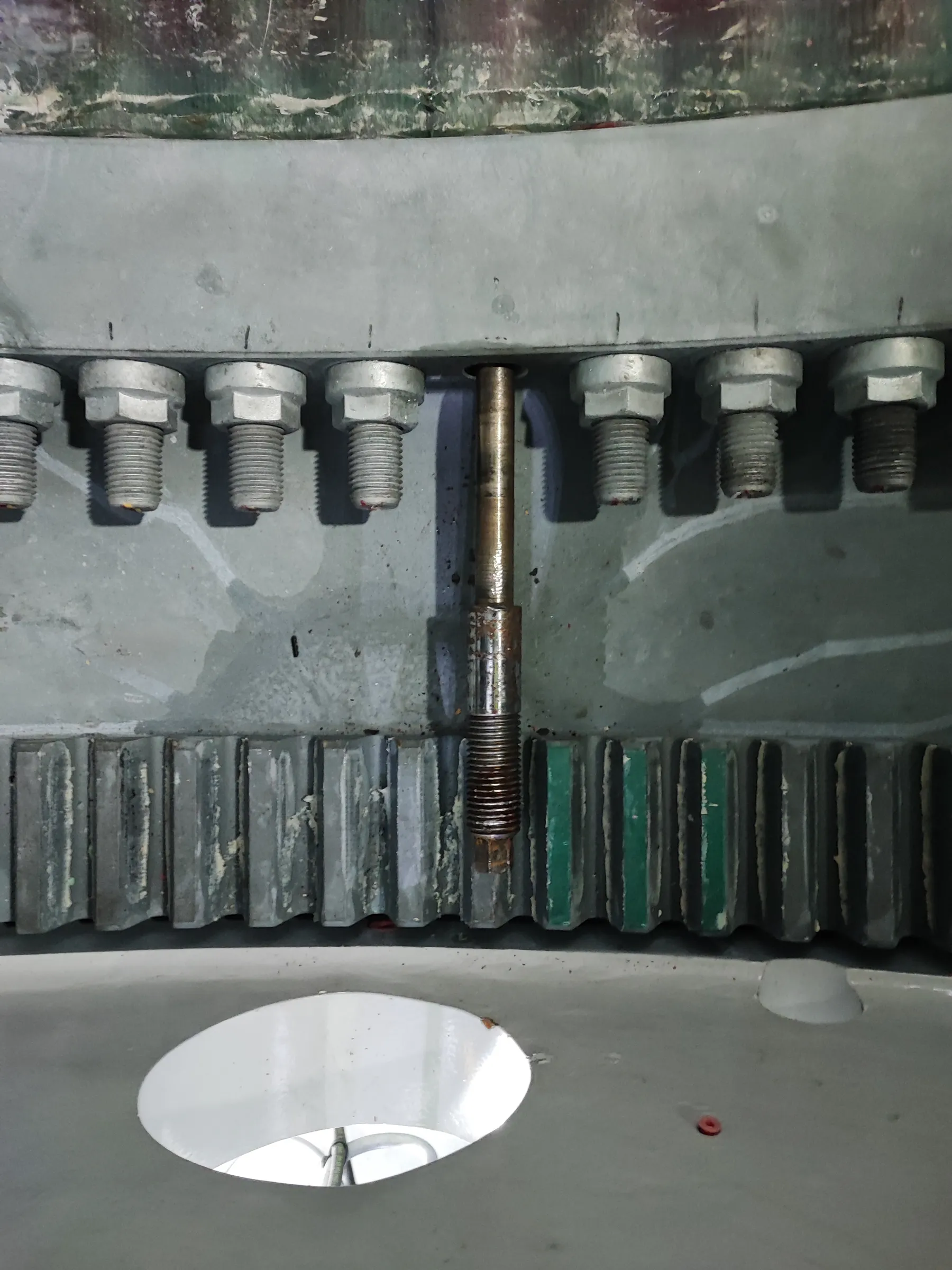 A close-up view of industrial machinery shows a grey metal beam with several silver bolts protruding downwards. A rusty threaded rod hangs centrally between two bolts. Below, a large grey gear or toothed belt features some teeth with white residue and others painted green. The bottom surface is light grey, with an oval opening revealing a bright white interior and a small red ring on the right.