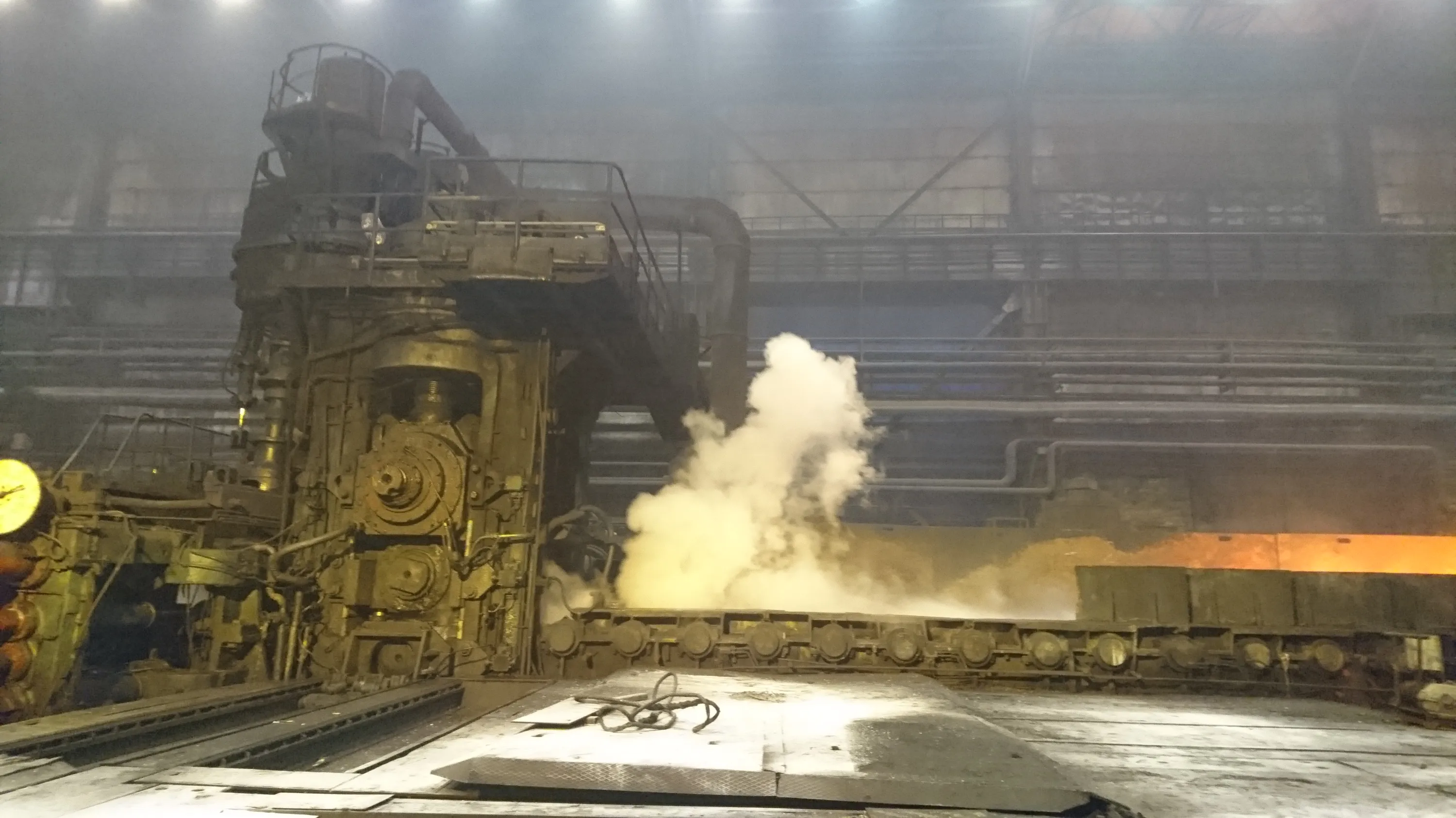 An active industrial facility features a massive, complex machine on the left, detailed with gears and pipes. A bright orange, glowing metal slab moves along a roller conveyor system on the right, emitting a large plume of white and yellowish smoke or steam. The vast, dimly lit interior is hazy, with overhead lights illuminating structural beams and elevated platforms in the background. The foreground shows a grimy floor with scattered cables.