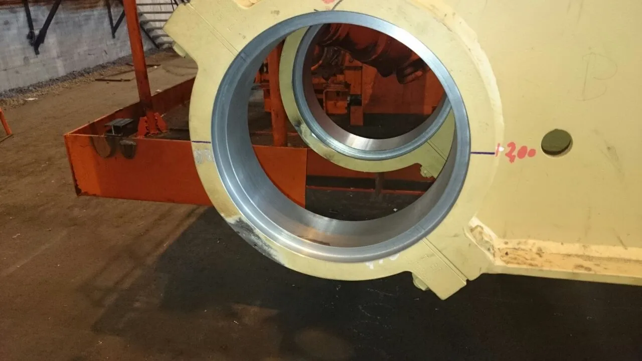 A large, light yellow industrial component features two prominent circular openings, each fitted with a metallic liner. An orange structure is visible through these openings in the background. Markings including a blue line, red '200', and a small circular hole are present on the yellow surface. The ground is dark, and the scene is dimly lit.