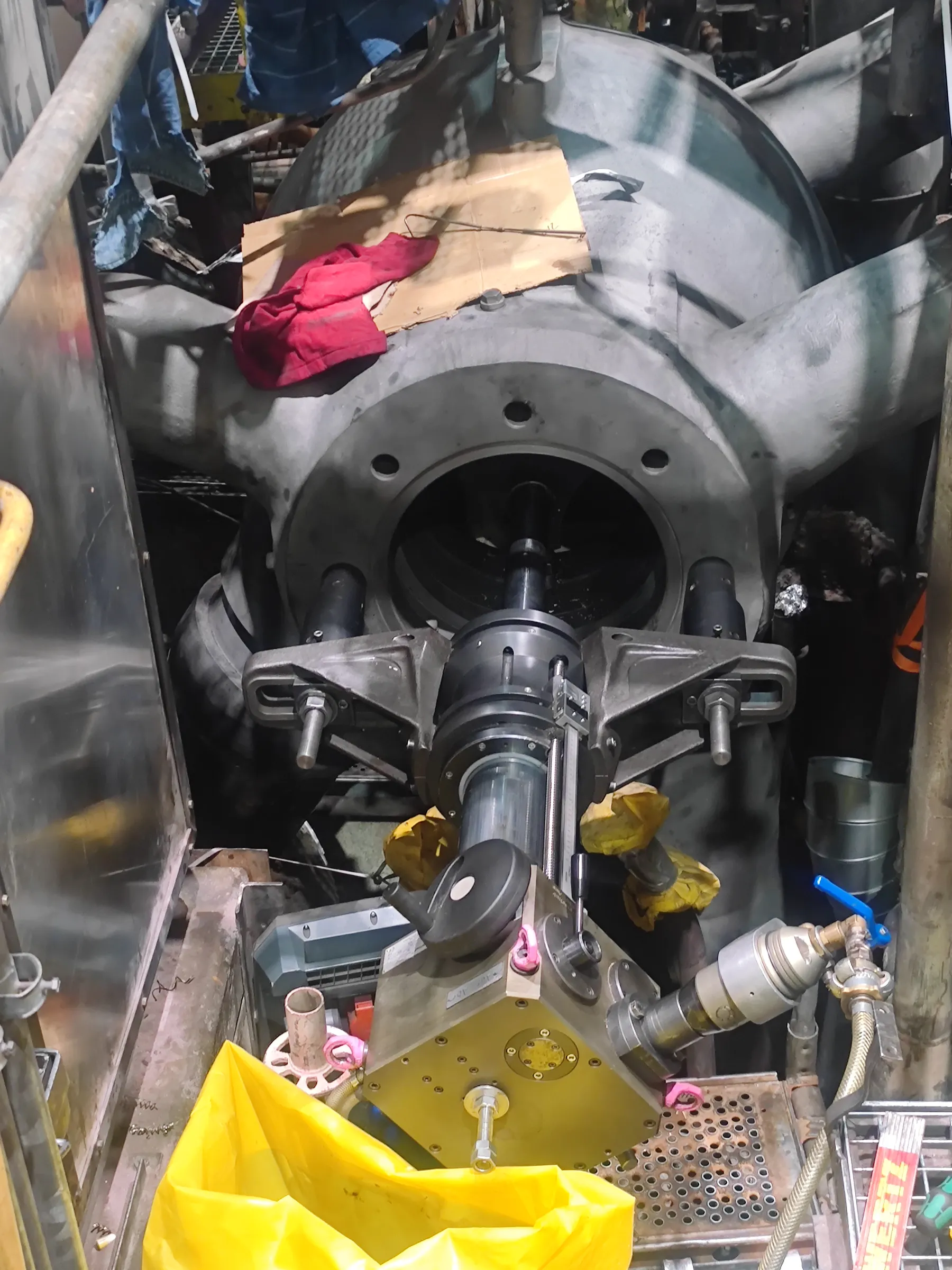 A large, grey industrial machine component, possibly a turbine, is shown with a specialized machining tool mounted in its central opening. The tool has black and grey parts, including a clear cylinder. Below, a metallic cube-shaped device with a blue-handled valve and braided hose sits near a bright yellow plastic bag, yellow gloves, and a red rag on cardboard. The scene is an active industrial workspace with artificial, somewhat dim lighting.