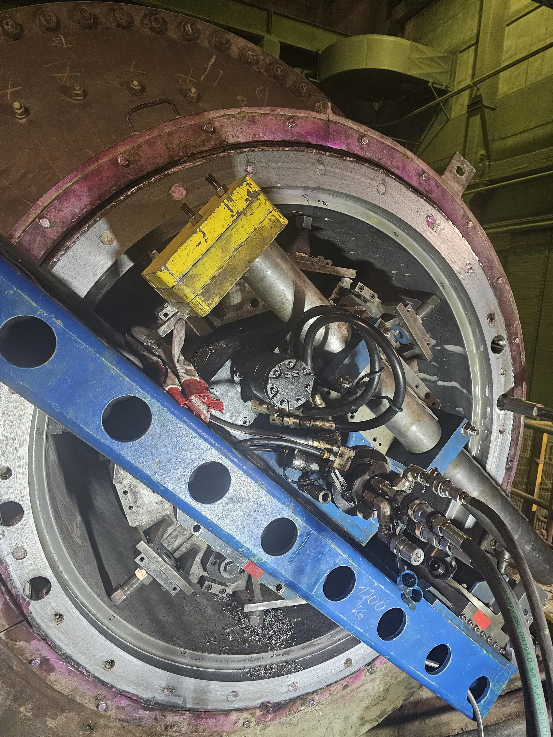 A close-up, angled view of a large industrial machine opening, revealing various tools and components. The outer reddish-brown flange, with a pinkish hue, is secured with numerous bolts, and an inner silver ring shows signs of machining. Inside the opening, a bright yellow block, a blue perforated beam, hydraulic hoses, a black HONEI pump, and metal shavings are visible. Work gloves and other metal fixtures are also present. The background shows olive-green industrial structures and ductwork under artificial lighting.