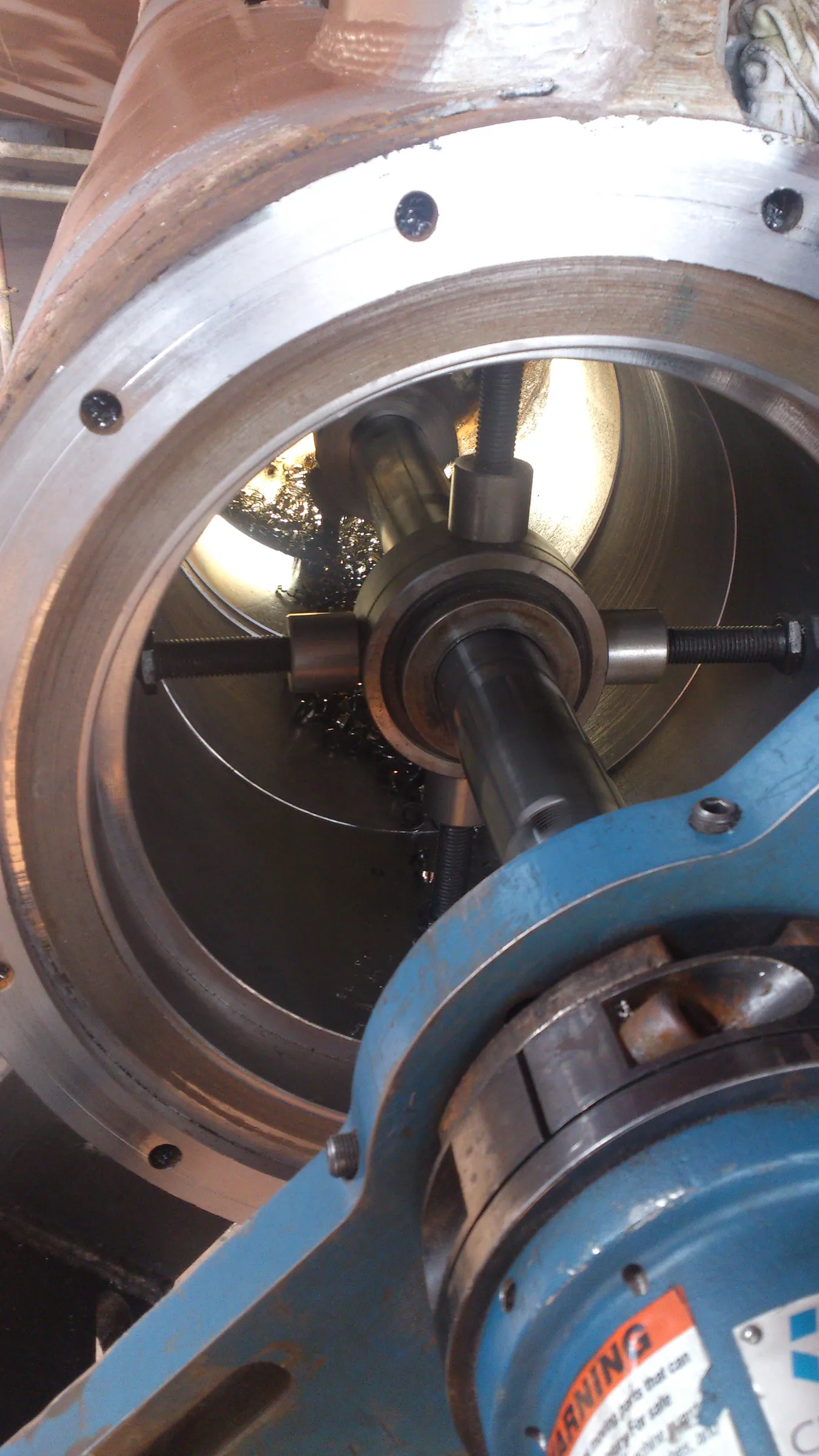 A close-up, top-down view shows an industrial boring machine operating inside a large, cylindrical metal housing. A central metallic shaft, part of the machining tool, extends into the bore, supported by adjustable rods pressing against the inner wall. Bright, curled metal shavings are scattered at the bottom of the cylinder, illuminated by internal light. The housing displays areas of dull brown and clean, bright silver metal. A dark blue machine component with a partially visible orange warning label is in the foreground.