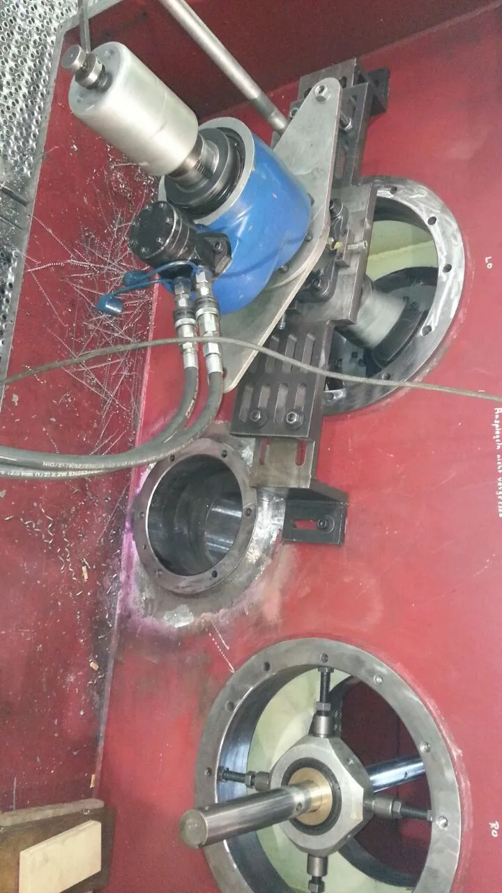 An industrial close-up shows a red machine housing with a blue and silver portable boring tool mounted on it. Silver metal shavings are scattered on the red surface, indicating recent machining. Black hydraulic hoses connect to the tool. Below, a newly machined circular bore is visible, and further down, another circular opening contains a central shaft supported by a four-spoke alignment fixture. Faint German text is written on the red surface.