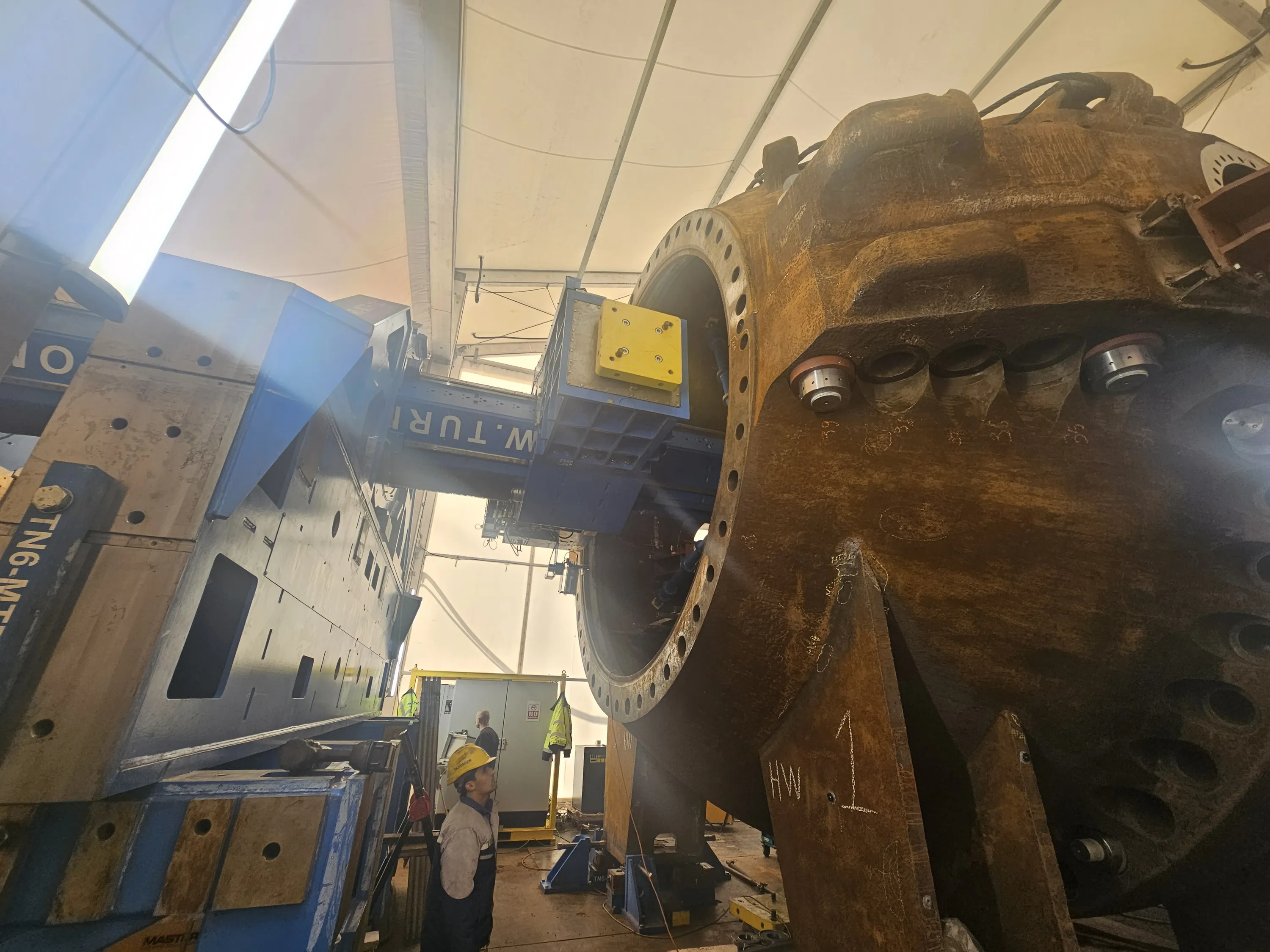 An industrial workshop scene shows a large, dark blue and grey machining tool positioned to work on a massive, rusty brown metal component. The cylindrical workpiece, marked 'HW 1' in white chalk, features a large flange with numerous bolt holes. A worker in a yellow hard hat and overalls stands in the foreground, looking up towards the machinery. Overhead, a white tent-like ceiling with bright strip lighting illuminates the space, revealing other equipment and a second worker in the background.