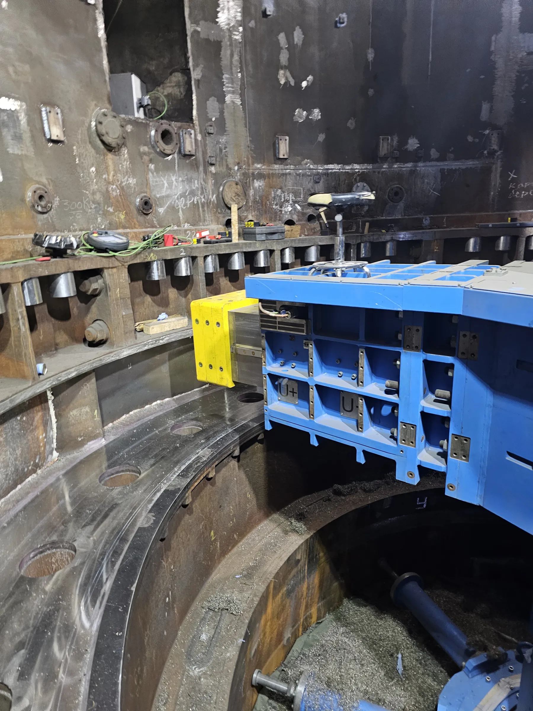 An industrial interior displays a large, dark grey and rusty metal structure, possibly a vessel, featuring various welded plates and circular flanges on its walls. A prominent blue machine with a bright yellow attachment is positioned over a large circular metal track or opening in the foreground. Metal shavings are scattered at the bottom right, indicating recent machining. Tools like a multimeter and clamps rest on a metal ledge above the track. The lighting is artificial and functional, illuminating the rough textures and construction details.