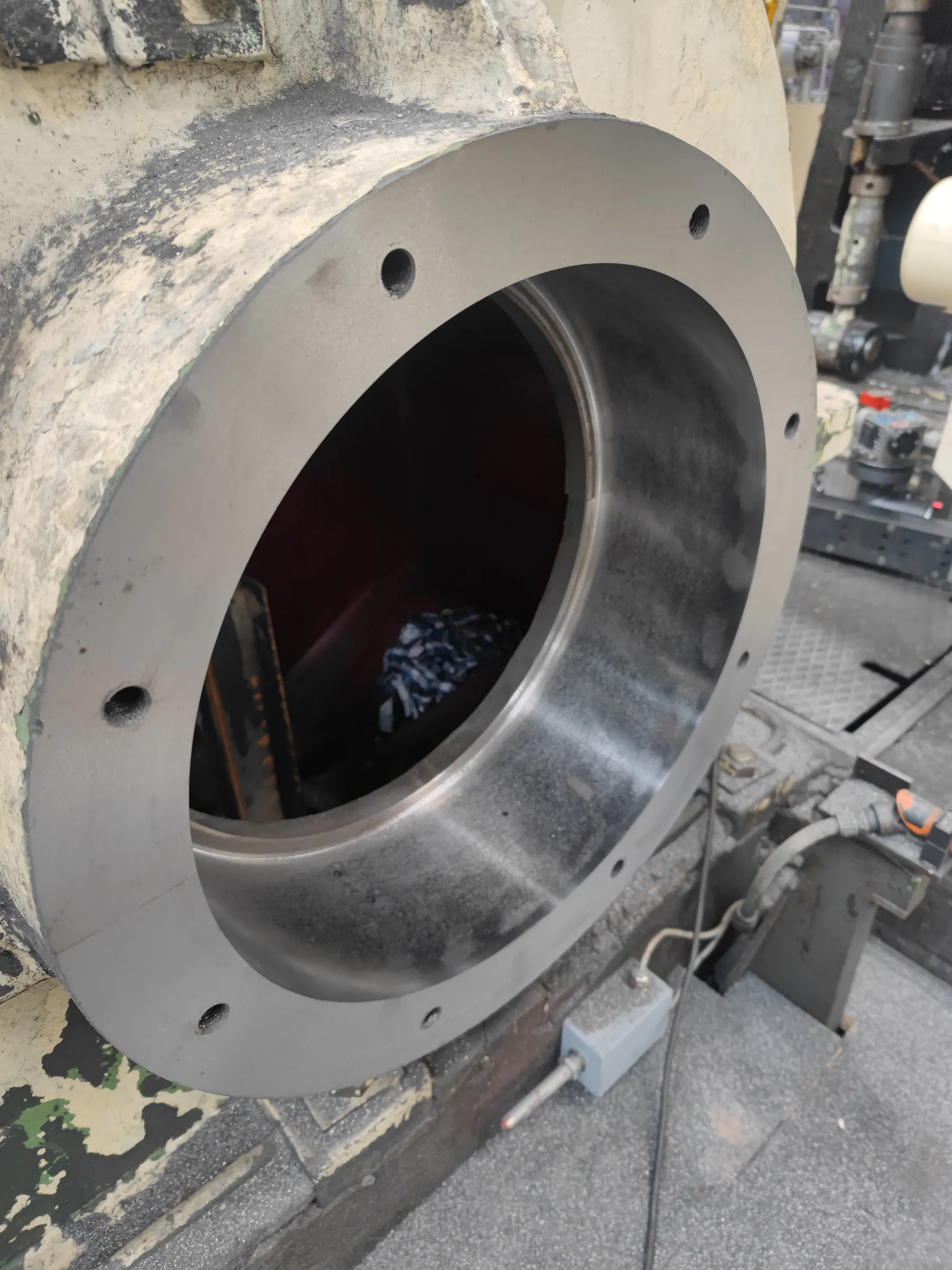 A large, industrial metal cylinder with a clean, grey flange and bolt holes is shown in a workshop. The cylinder's exterior is a grimy, light beige and dark grey, contrasting with the smooth, polished inner surface. Inside, a dark space reveals a patterned cloth. The background features other machinery, a textured grey floor, and a wired sensor, all under bright, functional lighting.