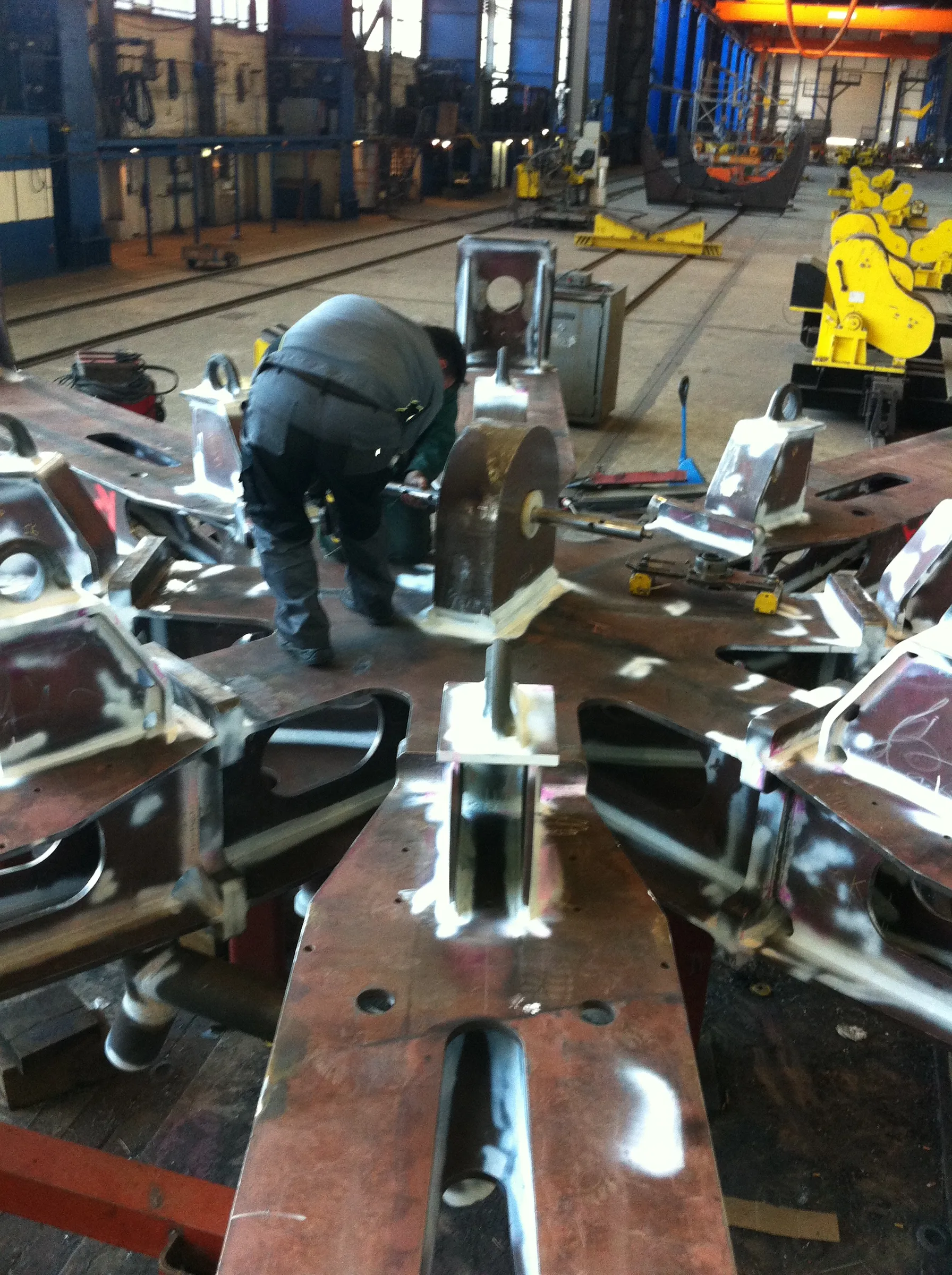 A worker in dark grey workwear is focused on a large, reddish-brown metal fabrication, which exhibits bright white ground areas, on a factory floor. The industrial setting includes floor tracks, yellow machinery, a substantial dark curved metal object, and blue structural supports under bright illumination.