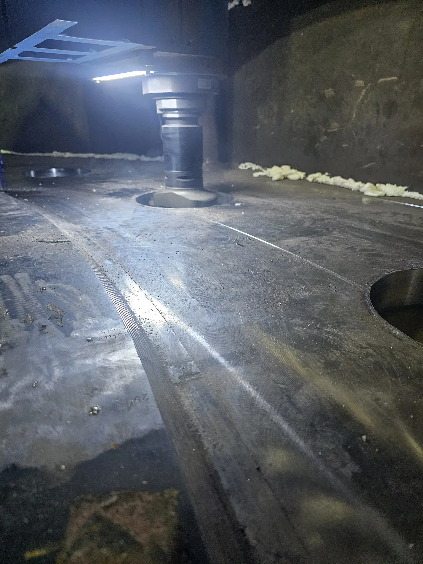 A large, dark metallic workpiece is being machined by a cylindrical tool, brightly illuminated by an overhead light. The surface displays machining marks, scattered debris, and some yellowish residue along the far edge.
