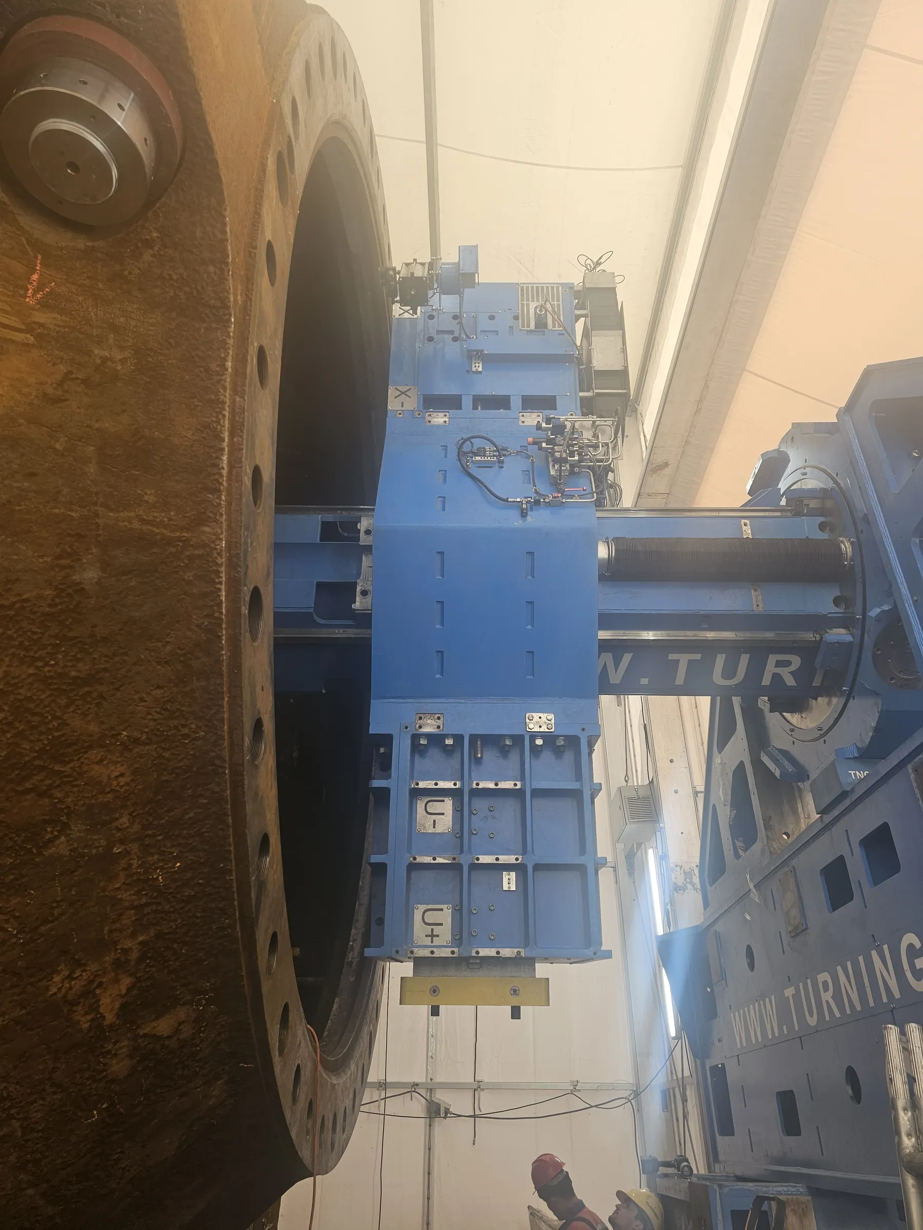A massive, rusty metal cylinder with a flanged end and bolt holes stands vertically, being machined by a large blue industrial turning machine. The machine, partially labeled "W. TURNING", features hydraulic lines, control panels, and a tool head extending into the cylinder's opening. The scene is brightly lit under a light-colored, tent-like ceiling, with a hazy atmosphere. Two workers in red and yellow hard hats are visible at the bottom right, observing the operation.