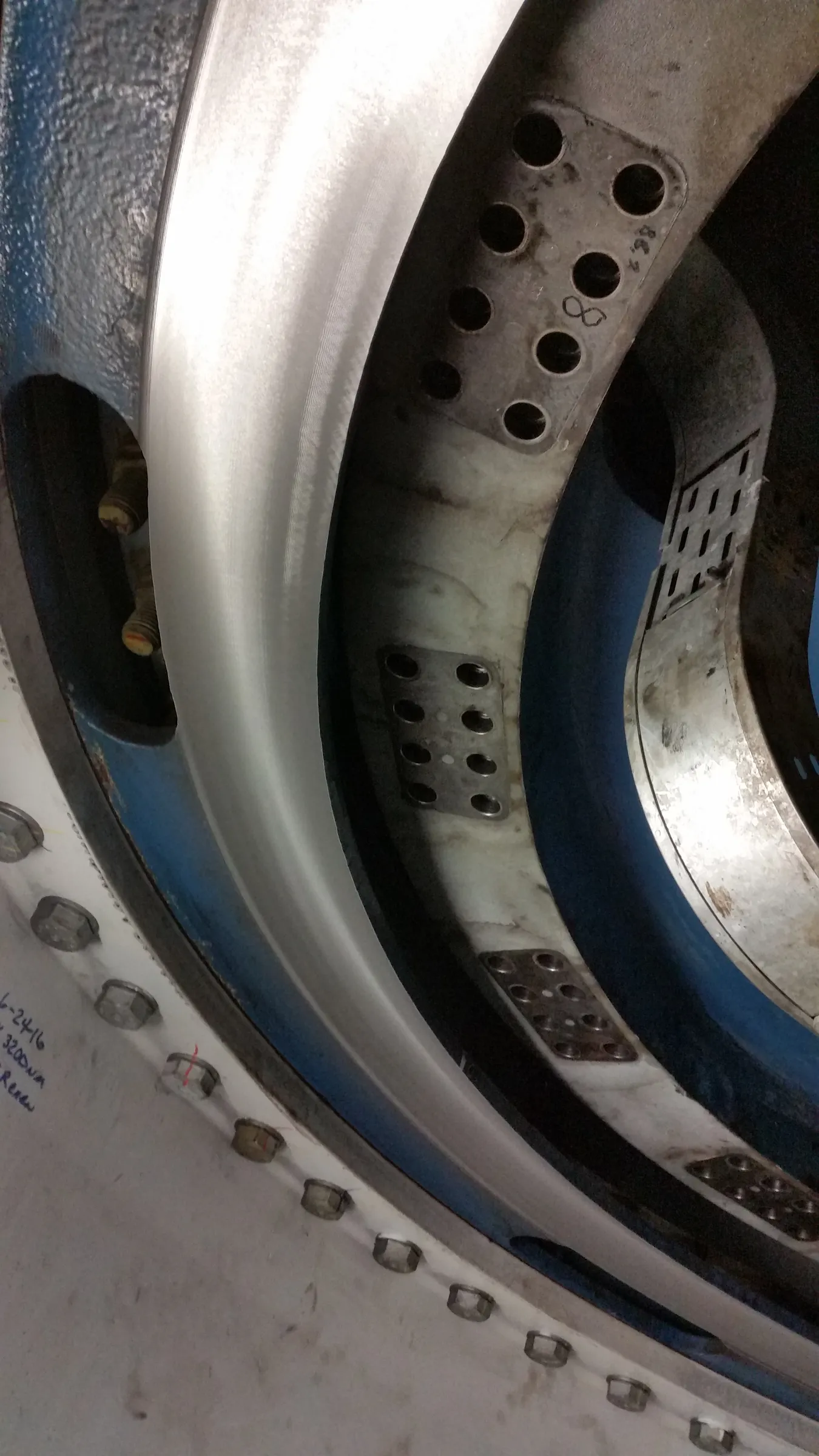 A close-up, angled view reveals multiple concentric industrial components, including a smooth, light grey curved metal surface with fine striations. To its left, a textured blue-grey surface shows two brass-colored cylindrical elements. To the right, a darker metal ring features rectangular plates with circular perforations, one marked with '∞' and '8C7'. In the foreground, a white surface with hexagonal bolts displays handwritten text: '6-24-16', '3200nm', 'Renew'.