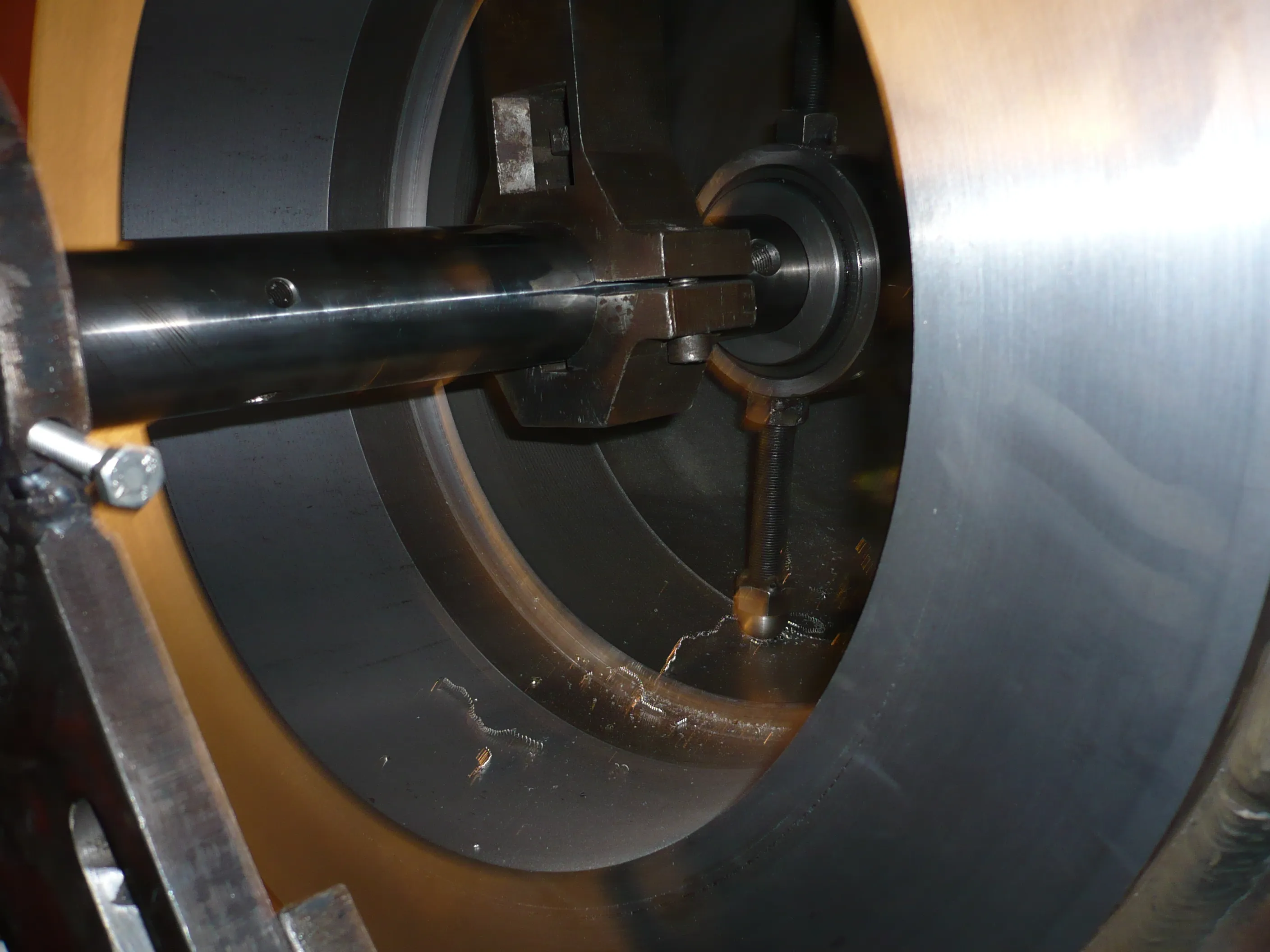 A close-up view of industrial machinery shows a shiny metal shaft extending through a dark gray cylindrical housing. A robust metal coupling connects to the shaft within the cylinder, where metal shavings are scattered on the bottom surface. Bright reflections illuminate the polished metal components and a silver bolt on a rough metal bracket, contrasting with the darker interior and a curved, yellowish-brown background.