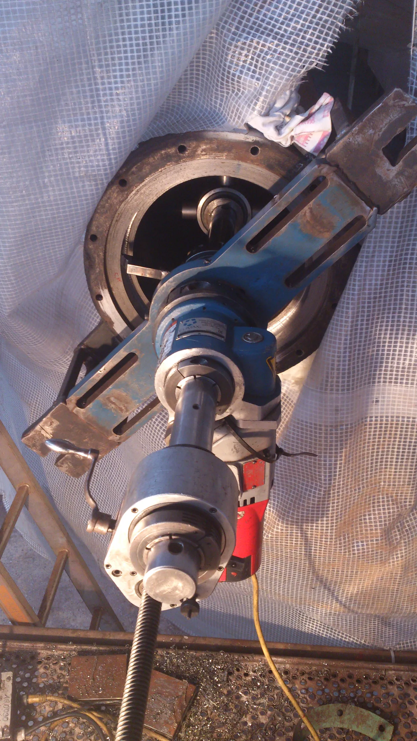 An angled close-up shows a blue and silver portable line boring machine attached to a large, rusty metal housing. A red power unit is connected to the machine, and a long threaded rod extends downwards. The background features a white, translucent grid-patterned fabric. Below the machinery, a metal grate is covered with numerous metal shavings, a yellow electrical cord, and other small metal pieces.