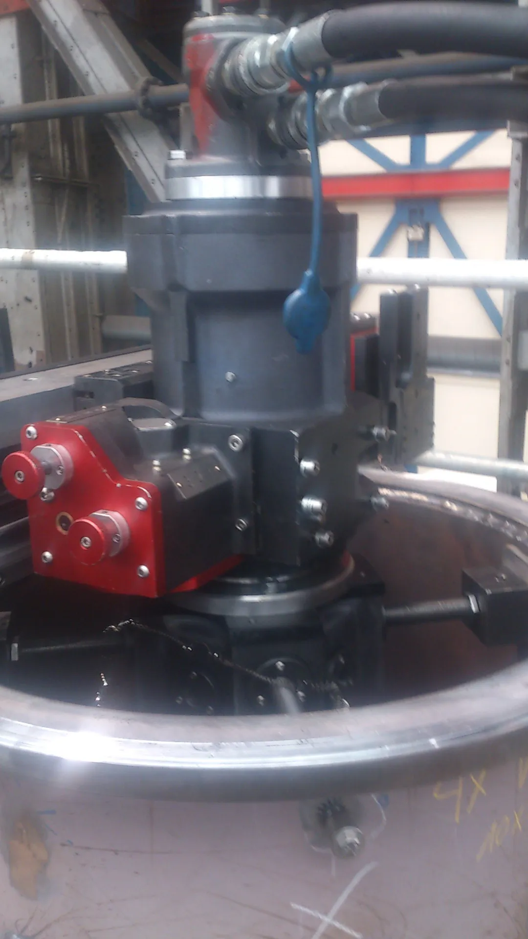 An industrial machine, featuring a grey and black main body, is positioned within a large reddish-brown metal cylinder. Hydraulic hoses connect to a red component at the machine's top, with a blue rubber object hanging from one hose. A red control unit with silver and red knobs is attached to the machine's side. Visible inside the cylinder are dark mechanical parts and a chain. The background shows grey metal structures and a blue and red truss, under industrial lighting.