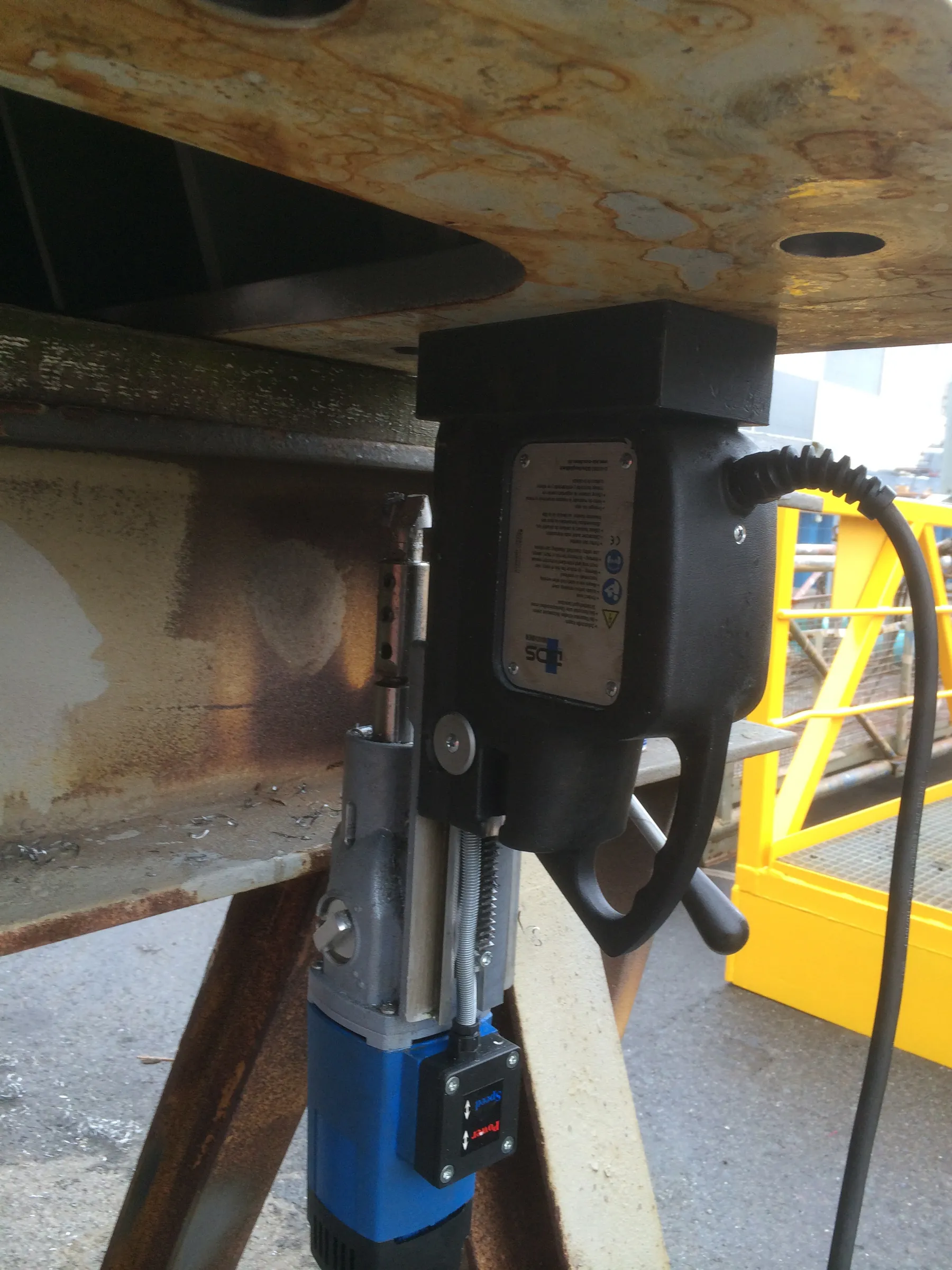 A magnetic drilling machine, primarily black and grey with a blue motor housing, is attached upside down to a large, rusty, light brown metal beam. A silver label with "TDS" branding and safety pictograms is affixed to the machine's side, from which a black power cable extends. Below the drill bit, silver metal shavings are visible on the lower beam. In the background, a bright yellow industrial structure with a grated platform is present, and the ground is dark grey. The scene is illuminated by natural daylight.