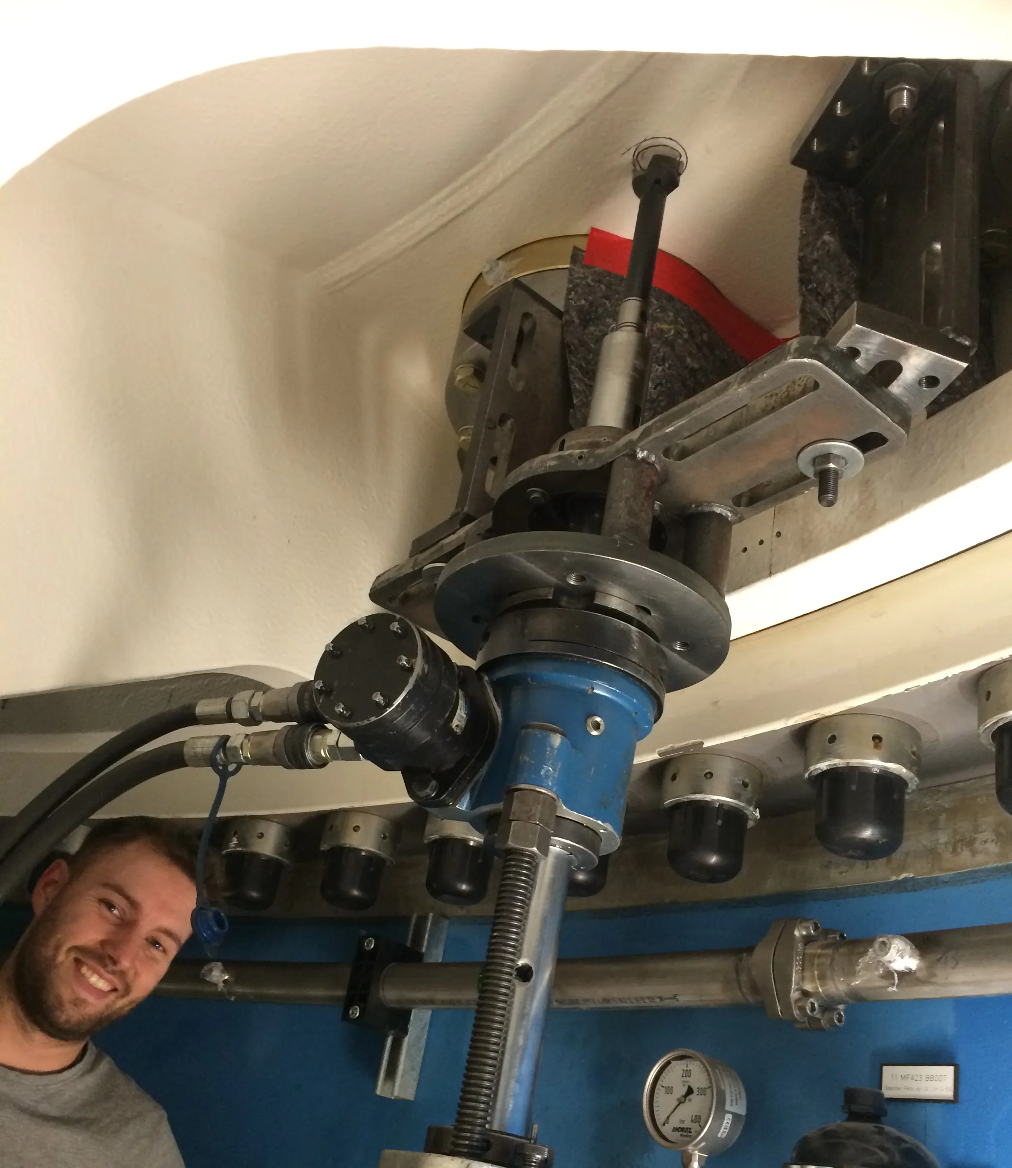 A man with a beard and grey shirt smiles from the lower left, looking towards the camera. Above him, a complex mechanical assembly is mounted against a white curved ceiling and a blue wall. The assembly features a prominent blue component with a large threaded rod, connected to various silver metal brackets and bolts. A black cylindrical unit with multiple screws is attached to the blue component, with two black hydraulic hoses leading from it, one secured by a blue cord. A row of black caps in silver metal rings is visible along a white beam. A horizontal metal pipe with a flanged connection is mounted on the blue wall, next to a pressure gauge displaying "MORITZ" and a range up to "400 bar", and a small black sign with white text.
