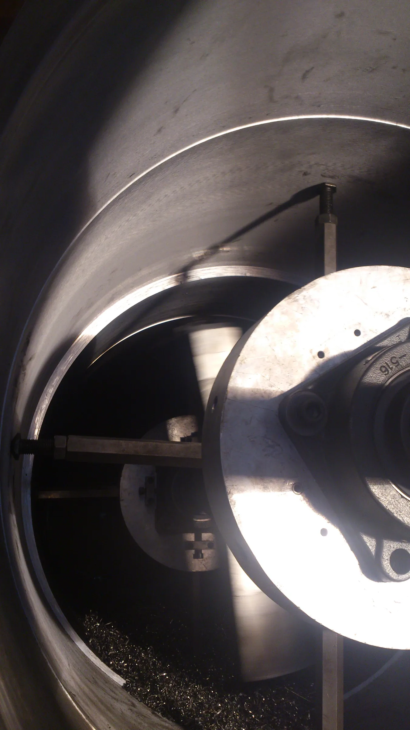 Inside a large metal cylinder, strong light highlights curved silver walls and a circular machine part with '516' markings. Metallic support rods cast long shadows over dark metal shavings collected at the bottom.