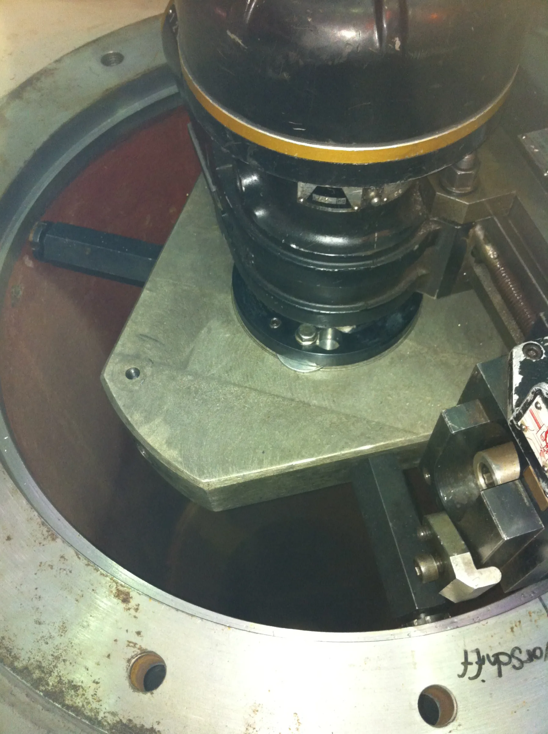 An overhead view shows a dark, cylindrical machine component with a yellow band, mounted on a light grey metal platform. This assembly sits within a circular, worn silver-grey metal casing, which has rust and handwritten German text "Vorschrift" on its edge. The interior background is a dark reddish-brown, with other metallic mechanical parts visible to the right.