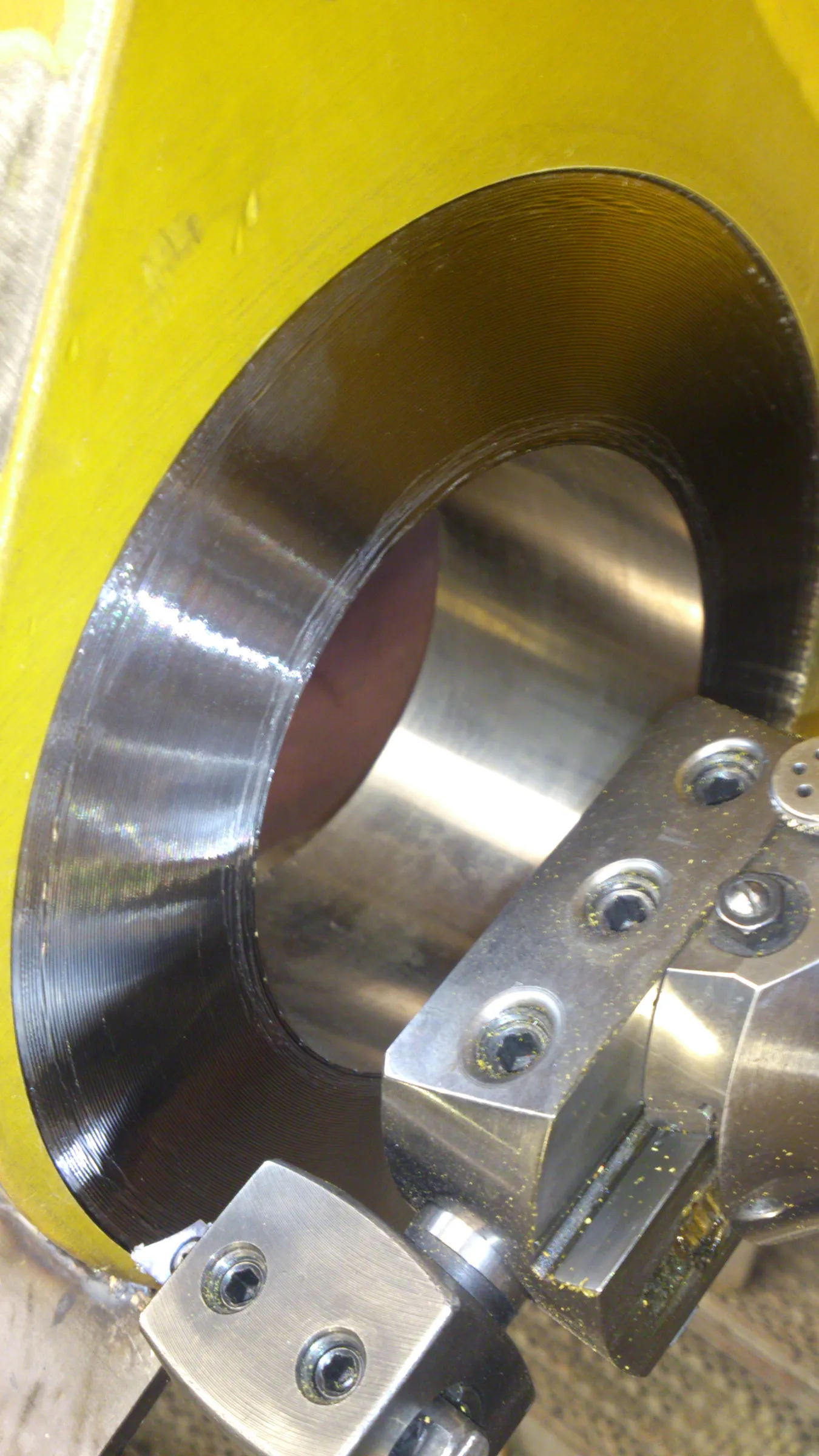 A close-up, high-angle view shows a metal machining operation. A large, cylindrical metal workpiece features a highly reflective silver interior and a dark, grooved outer surface. A metallic cutting tool holder, speckled with small yellow metal shavings, is positioned in the foreground, with a cutting insert visible near the workpiece. The bright yellow machine frame is visible on the left and top, providing a strong color contrast. The lighting highlights the reflections on the machined surfaces.