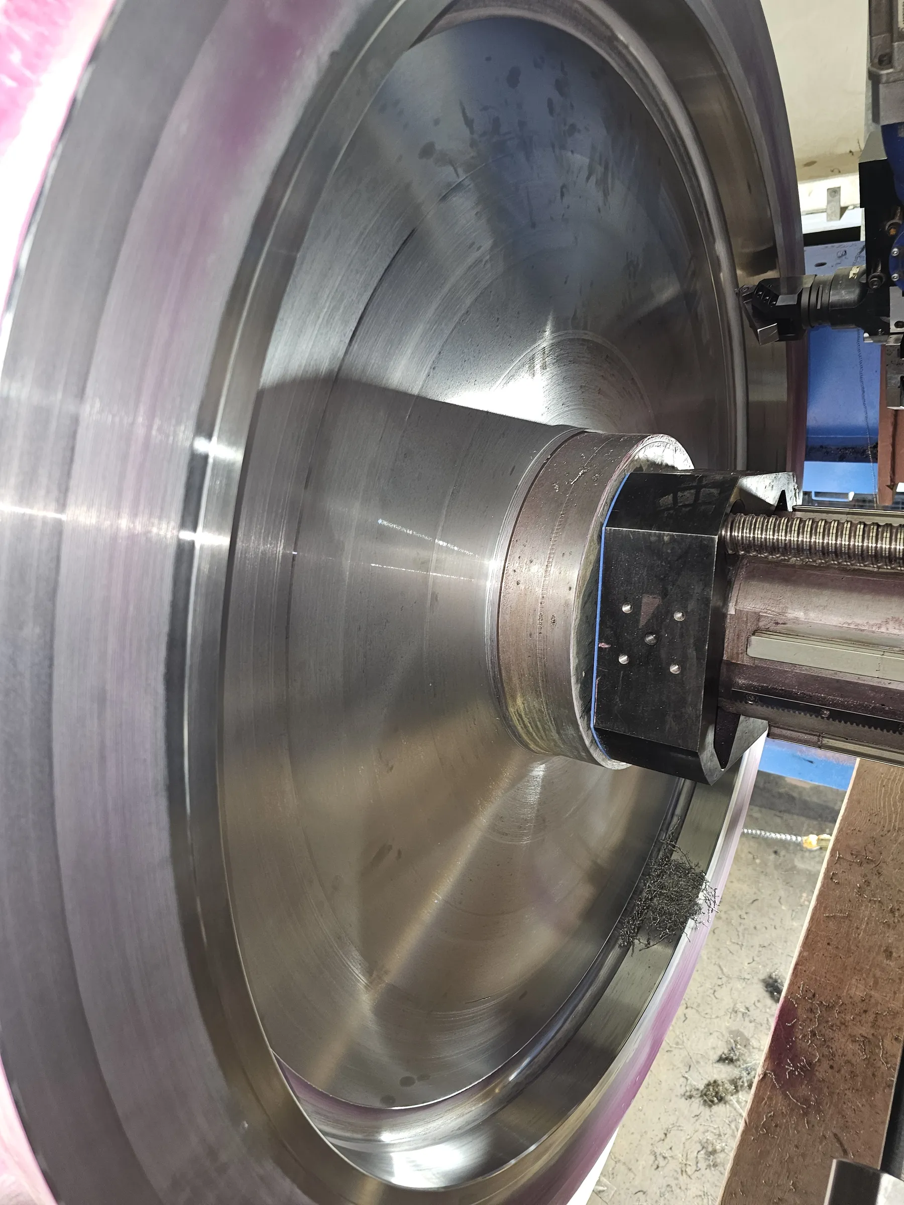 A large, circular metal component is being machined, with a cutting tool engaged near its outer edge. The metallic surface of the workpiece is shiny, reflecting light and showing concentric machining marks, with a faint purple tint on its rim. Metal shavings are visible below the cutting area. A threaded rod and a black linear guide block are part of the machine's mechanism on the right.