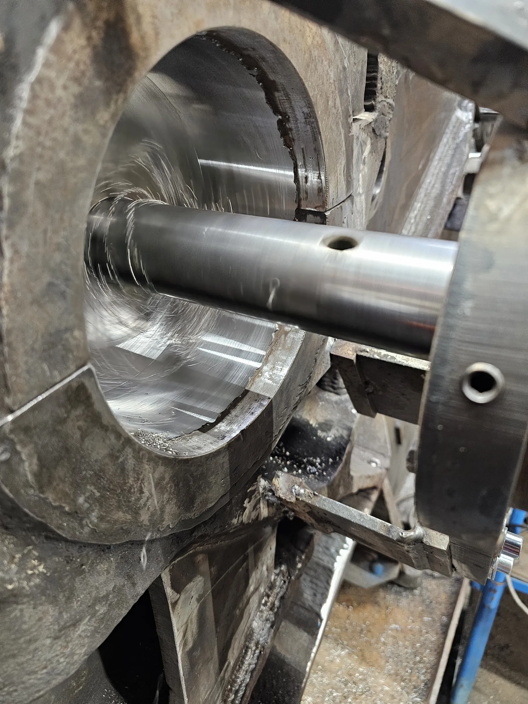 A close-up, vertical shot captures a large, cylindrical metal workpiece being machined. A bright, polished metal rod, likely a boring bar, extends horizontally through the center, surrounded by a flurry of fine, silvery metal shavings from the active cutting. The interior surface of the cylinder is smooth and reflective with visible machining marks, while the exterior and surrounding machine parts are rougher, dark gray metal. A small hole is visible on the metal rod. The scene is brightly lit, emphasizing the metallic textures and the ongoing industrial process.