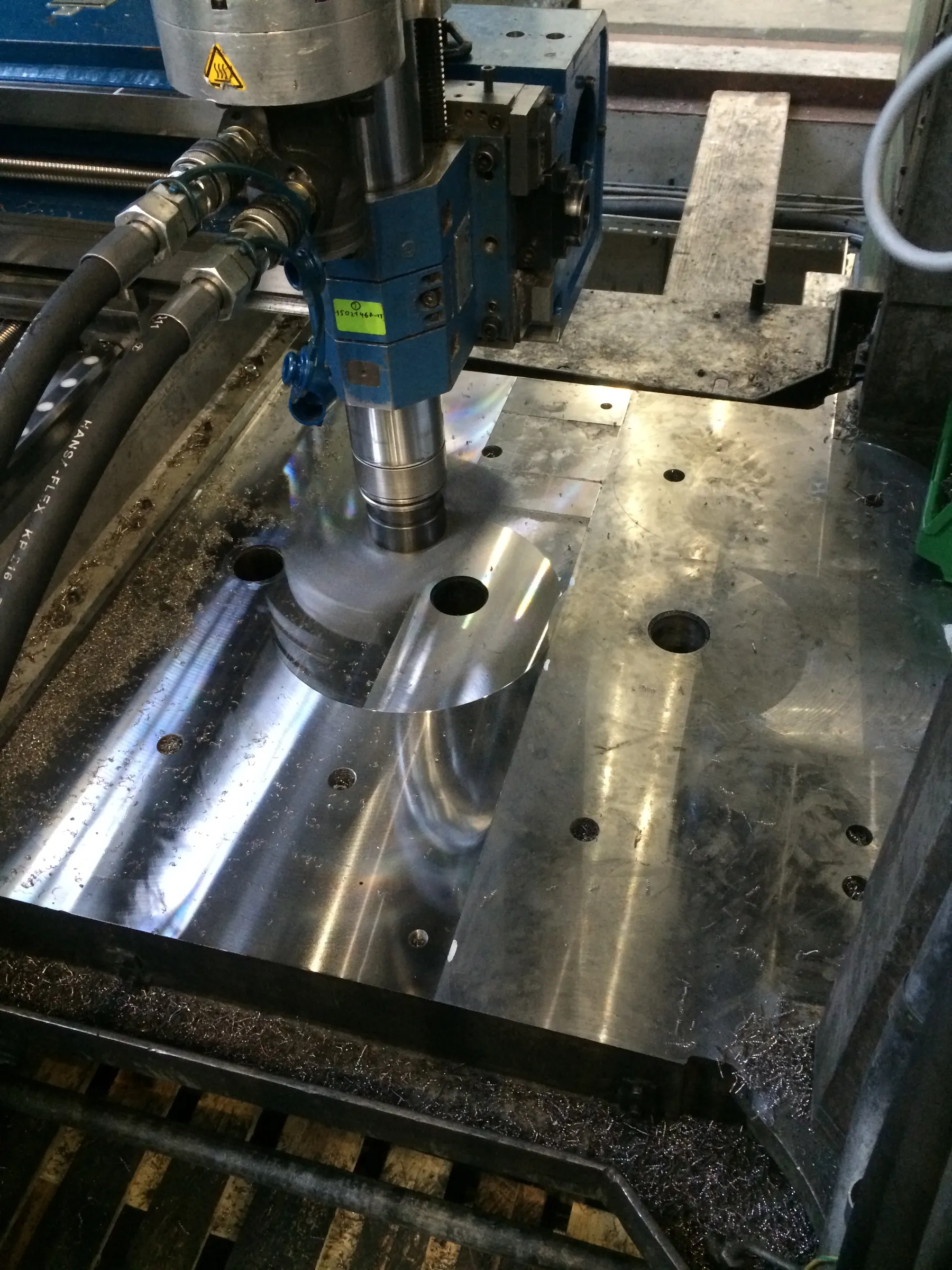A close-up view shows a blue and silver industrial machine actively milling a large, reflective metal plate. A cylindrical silver tool is engaged with the workpiece, creating a circular cut, while abundant silver metal shavings are scattered across the surface and collected in a dark trough below. Black hydraulic hoses with silver fittings connect to the machine's head, which displays a yellow hot surface warning symbol and a green identification label. Bright lighting creates strong reflections and iridescent highlights on the freshly machined metal.