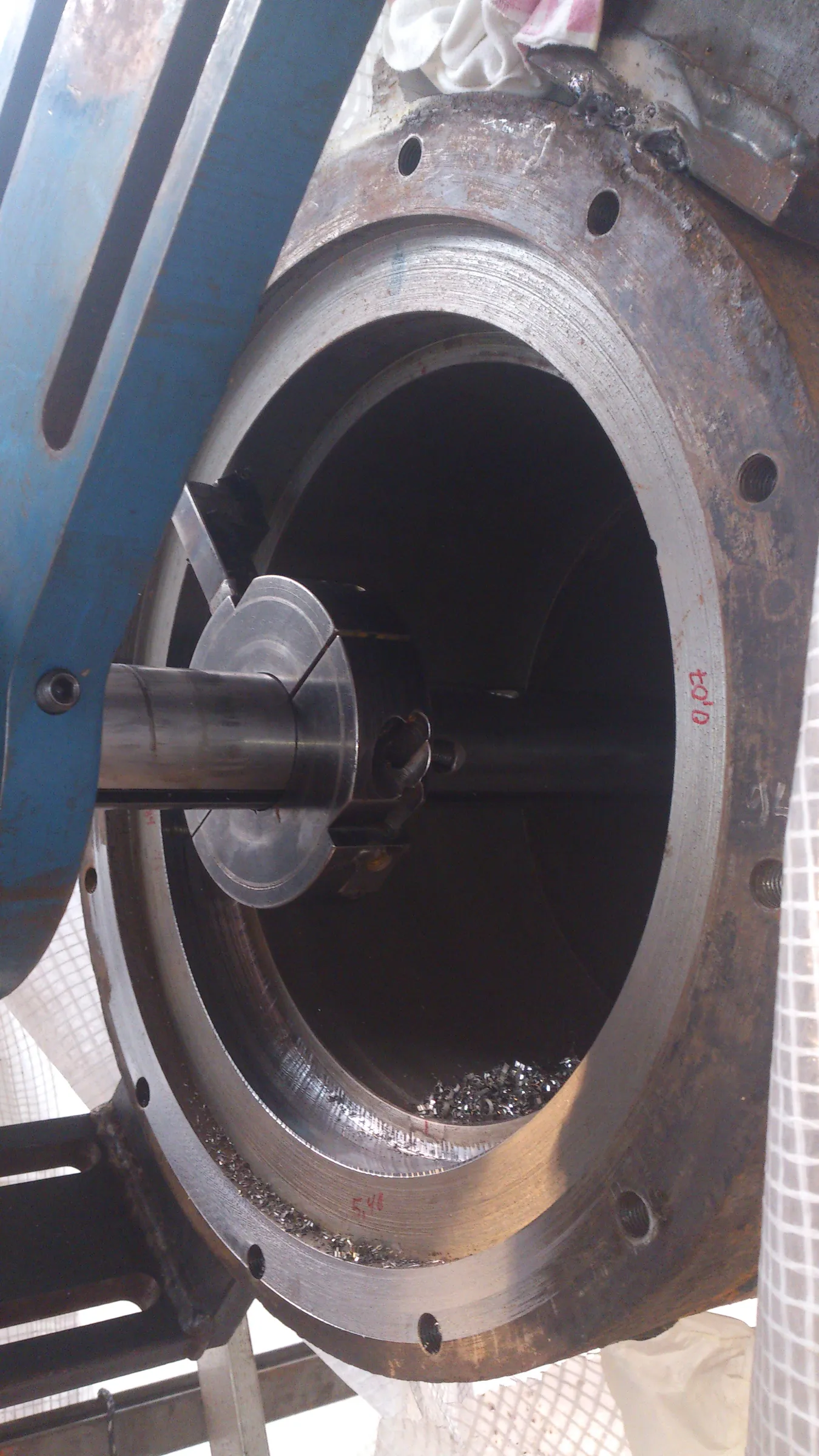 A large, circular metal opening, likely a pipe or machine component, is shown undergoing machining. A blue metal arm supports a central shaft with a cutting tool inside the dark interior. Shiny metal shavings accumulate at the bottom. The outer flange is rusty brown with bolt holes and red handwritten numbers. A white and red striped cloth is visible at the top right, near some rough welding, and a white mesh material is on the far right.