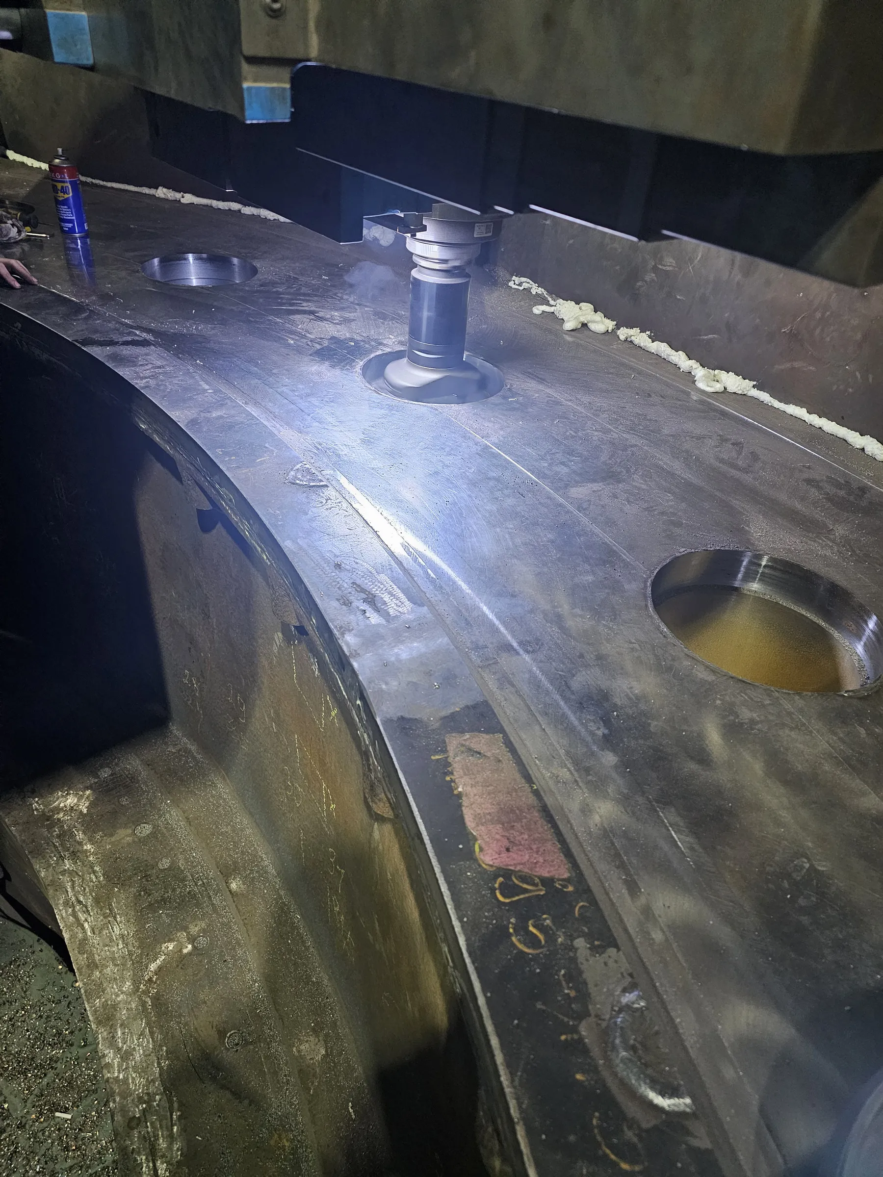 A large, dark gray industrial metal component is undergoing a machining process, with a rotating cutting tool actively boring into a circular opening. Intense light illuminates the immediate work area, reflecting brightly off the metal surface and the tool. To the left, a finished circular hole and a blue can of WD-40 are present, with a person's hand partially visible. A line of white, foamy material runs along an upper edge, and the curved side of the component displays various markings and textures.