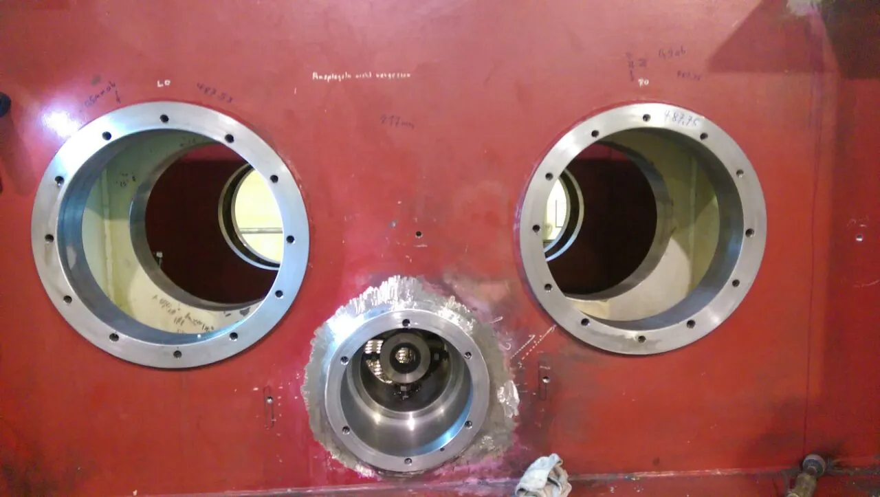 A large, reddish-brown metal panel displays three circular openings. Two prominent circular ports, one on the left and one on the right, are fitted with bolted silver metallic flanges. A smaller, central circular port at the bottom also has a metallic flange and contains visible internal machinery. Rough, silvery material surrounds the central port. Faint white handwritten text and numerical markings are present on the panel's surface. The scene is brightly and evenly lit, with reflections on the metal and light visible within the openings.